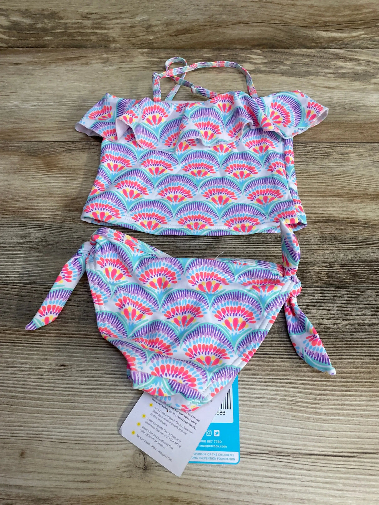 Flat-lay of 2T tankini with ruffled top, crossed straps and side-tie bottoms in pink, purple and teal scallop print on wood
