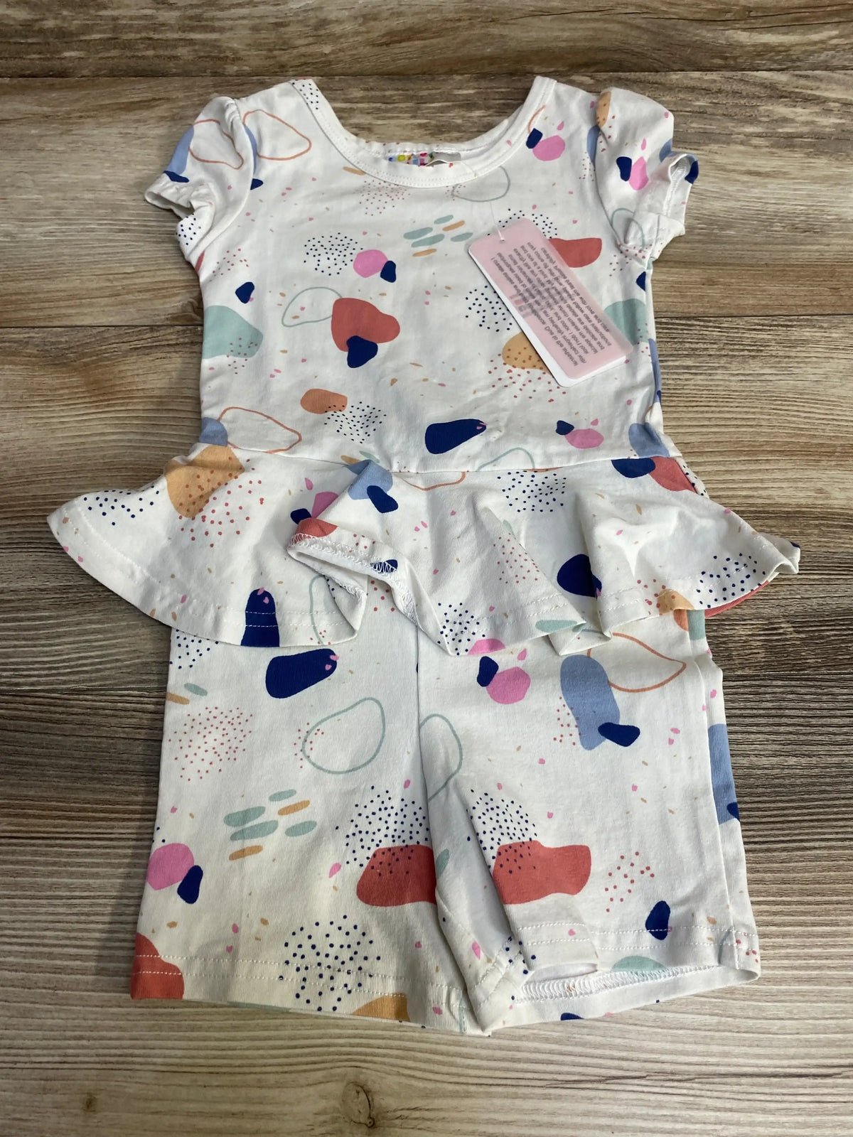 Flat lay of white baby romper (size 12-24m) showcasing multicolor terrazzo print, peplum waist and short sleeves on wood background