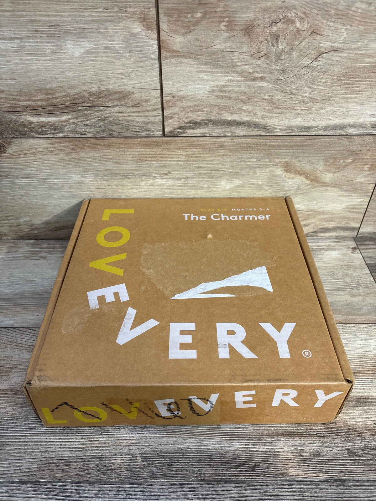 Front view of Lovevery The Charmer Play Kit box labeled Months 3-4 with wood-grain floor background