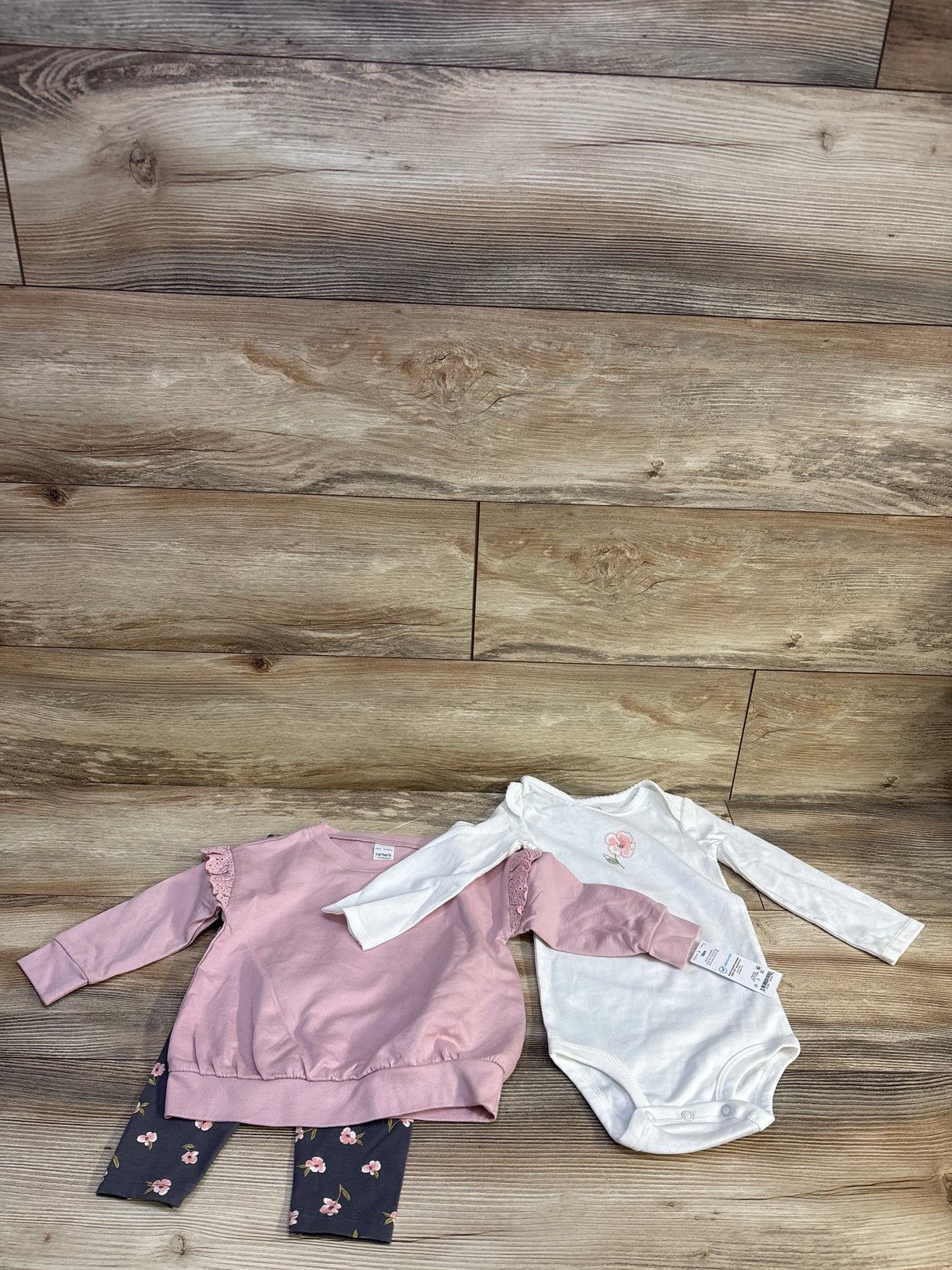 Flat lay of 3-piece baby floral set showing pink ruffle-sleeve sweatshirt, navy floral leggings, and white long-sleeve bodysuit on wood background