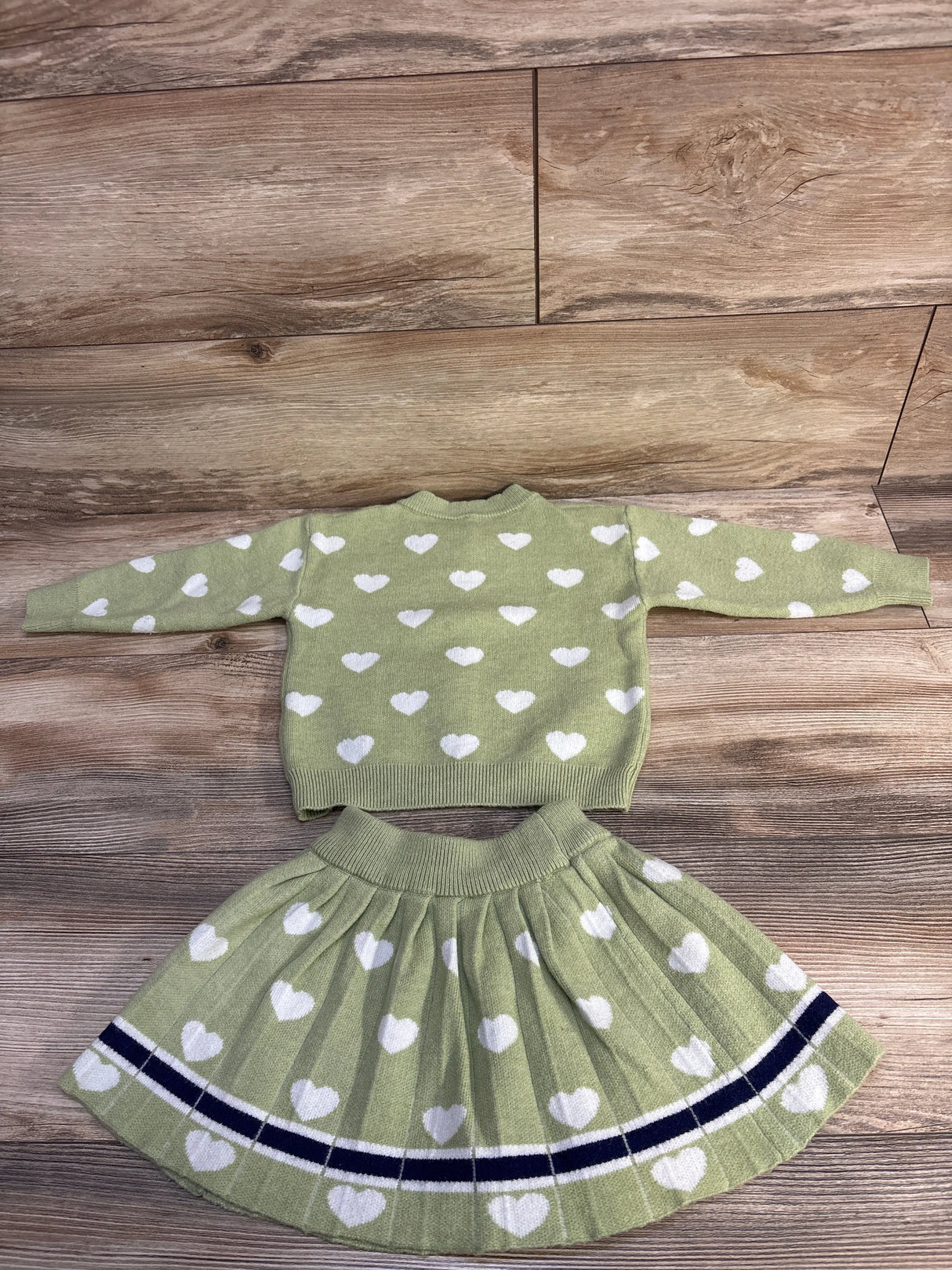 Green toddler sweater and pleated skirt set with white heart patterns on wooden floor