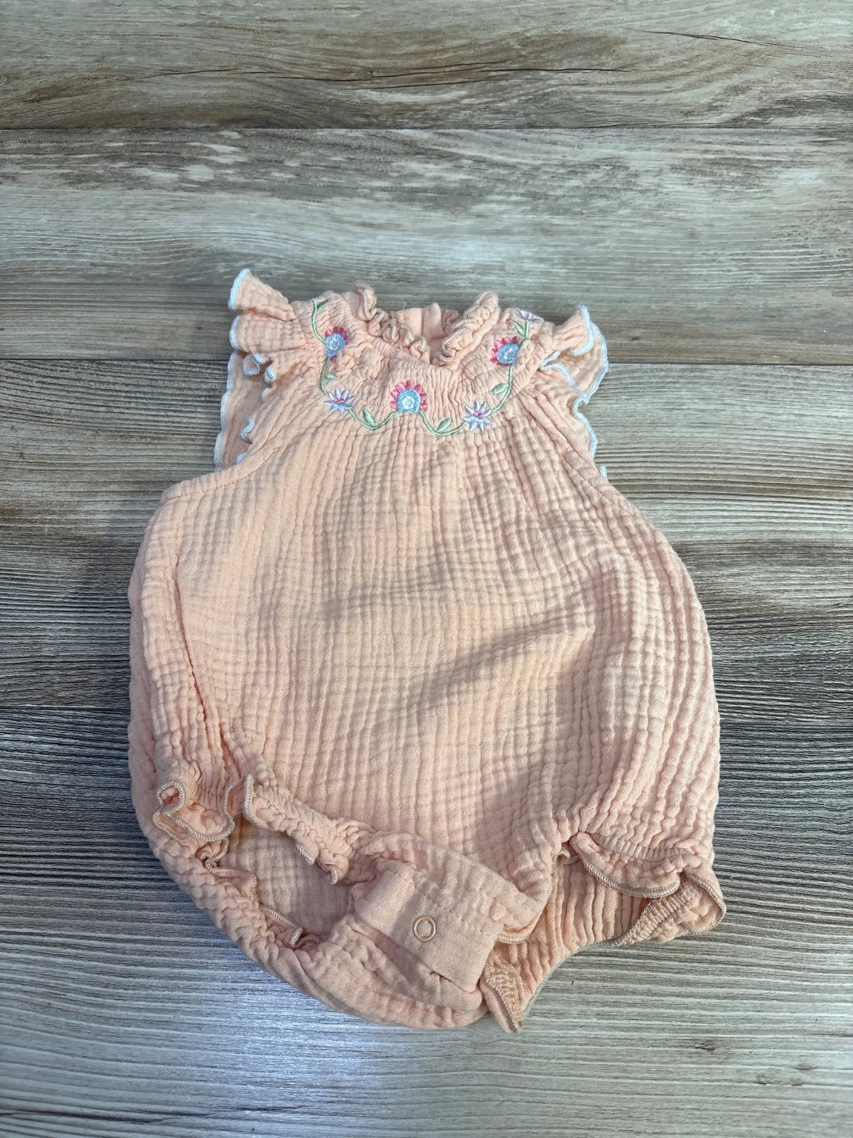 Top-down view of peach smocked romper (3-6 months) with embroidered flowers, ruffle sleeves and crotch snaps on wood