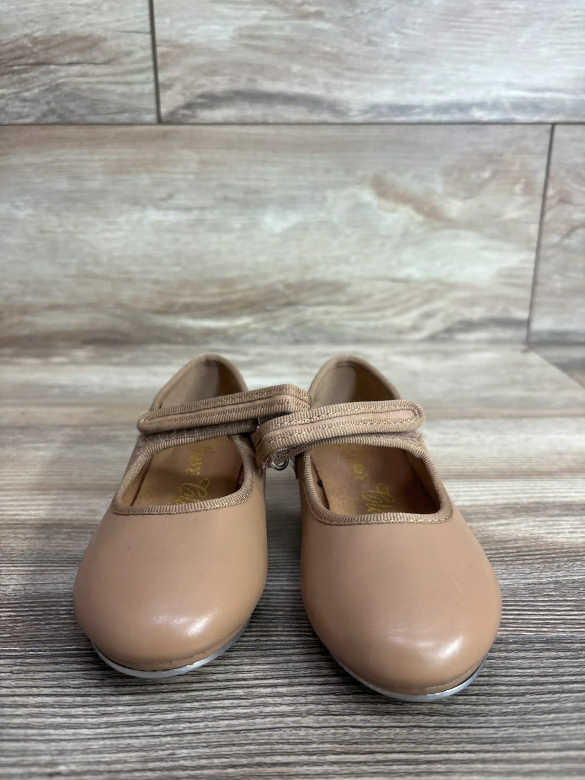 Front view of tan toddler tap shoes showcasing Mary Jane velcro strap and metal toe taps with wood plank background