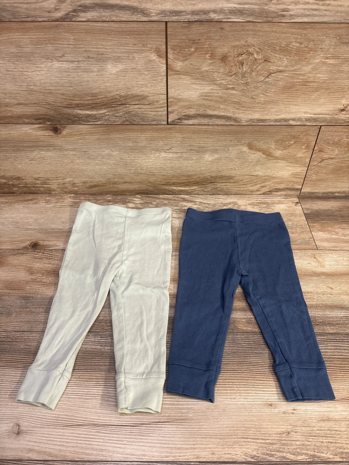 Overhead view of Burt’s Bees Baby 2pk pull-on pants in blue and light gray, showcasing elastic waists and cuffs on a wood-look floor