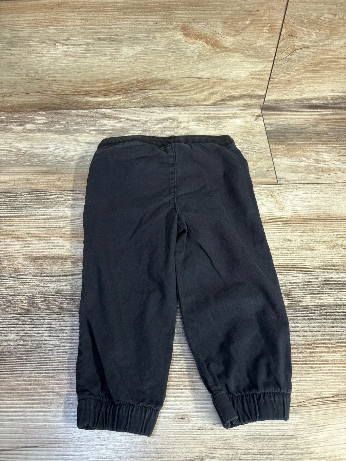 Front view of black Jumping Beans drawstring joggers in size 18 months showcasing elastic ankle cuffs on a wood-look floor