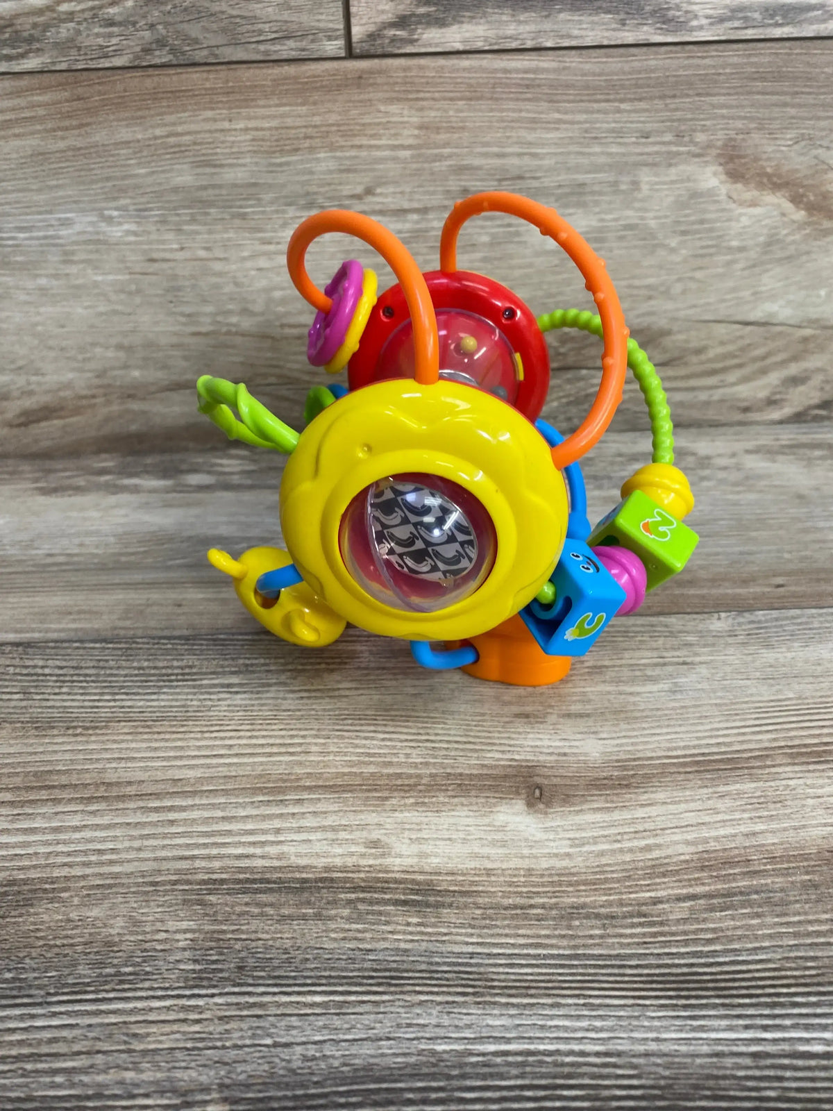 Angled view of Grab N Shake sensory ball showing yellow spinning rattle, orange loops and teether on wood background
