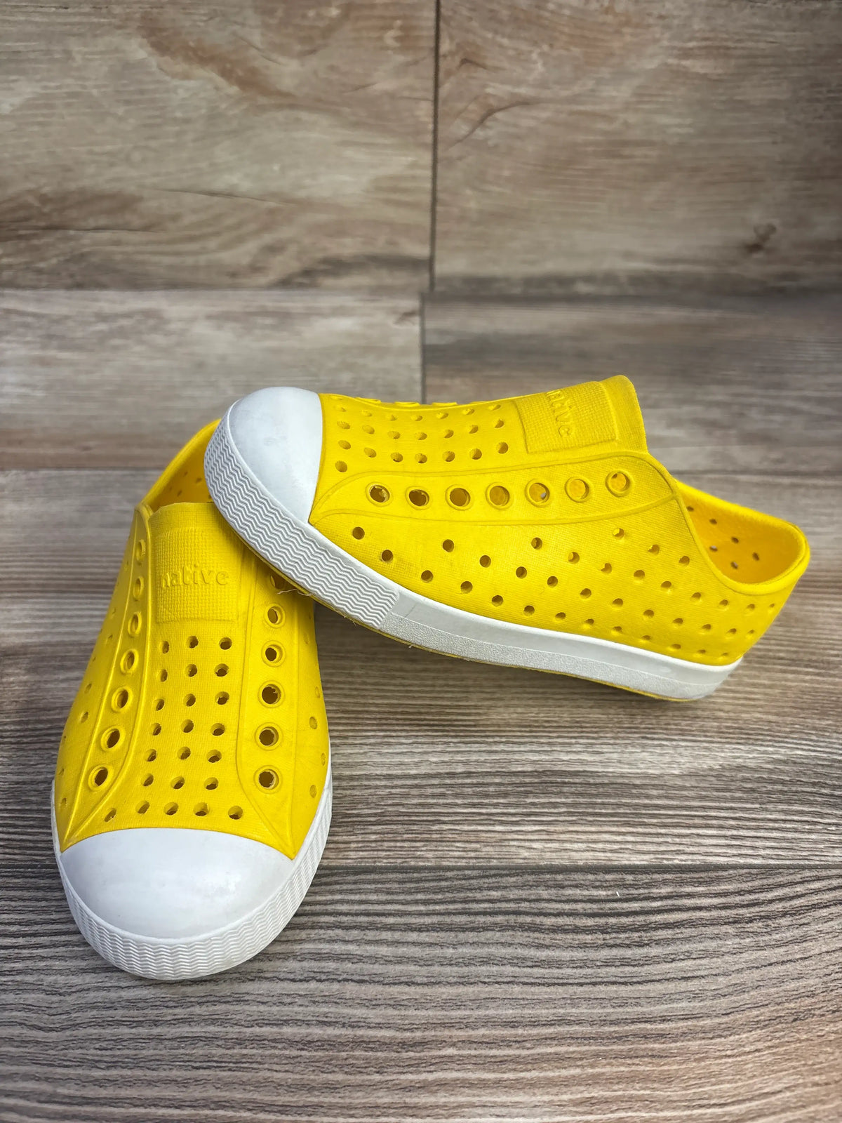 Angled view of yellow Native Jefferson slip-on shoes showcasing perforated uppers and white toe caps on a wood-grain surface