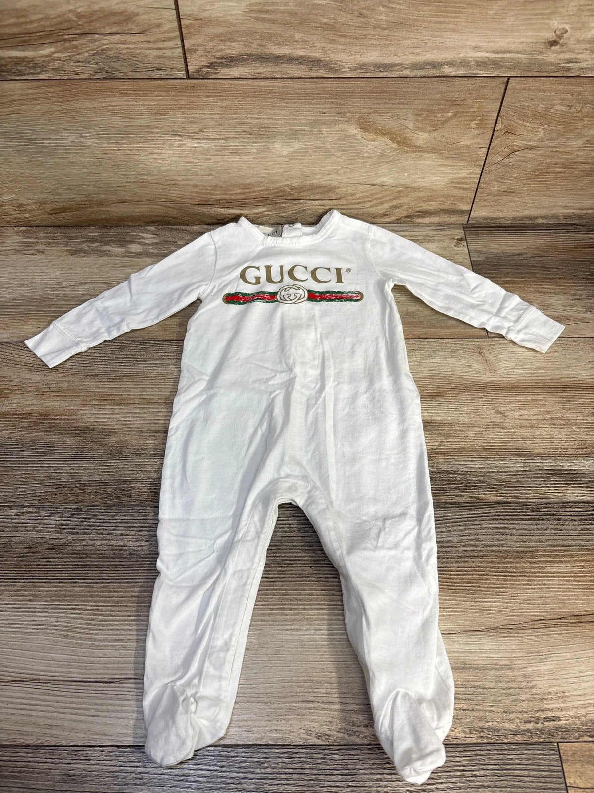Front view of Gucci white cotton logo footie sleeper, size 6/9 months, with long sleeves and enclosed feet on a wood floor