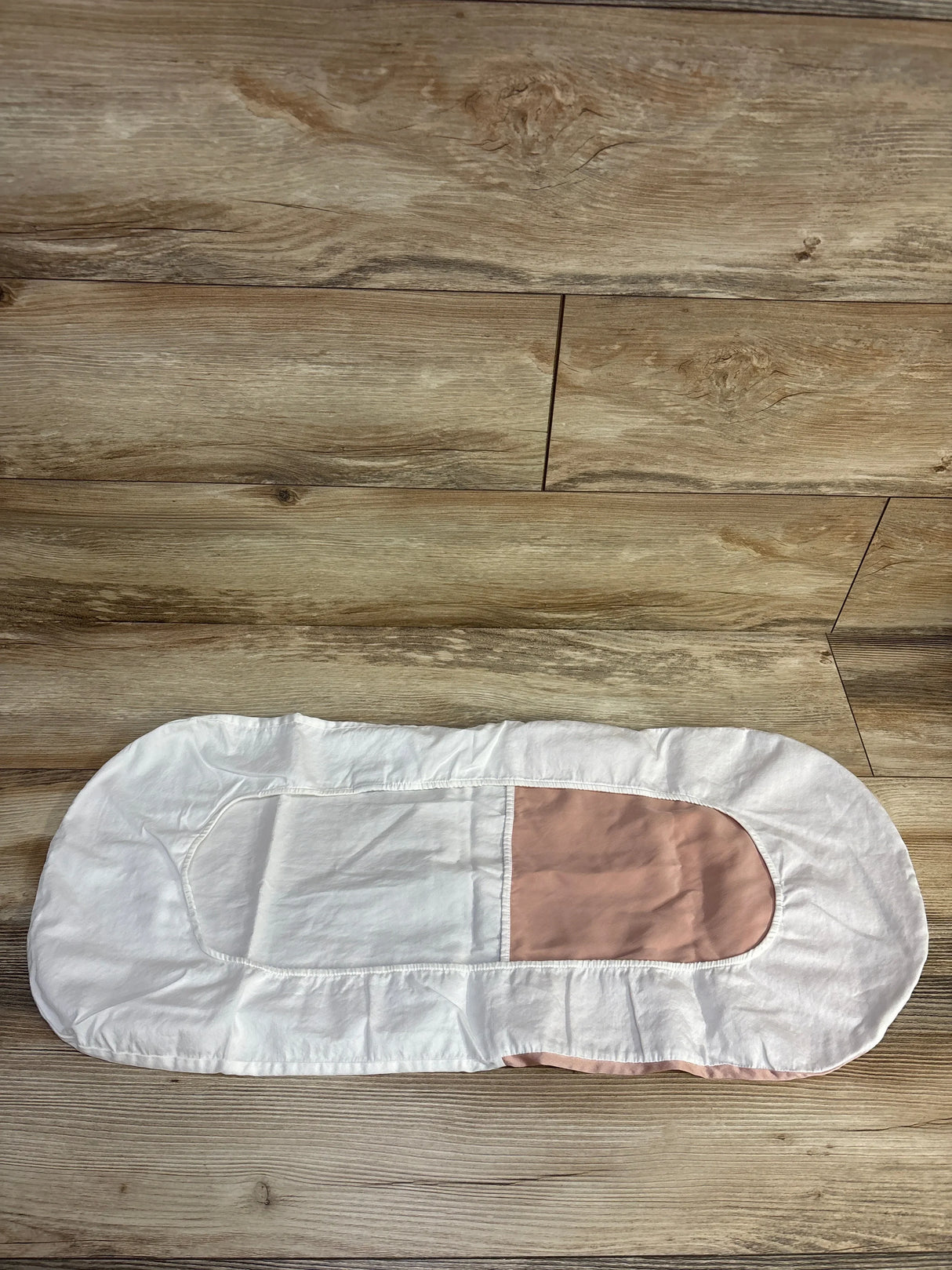 Top view of Silky Tots Silk oval bassinet sheet in pink and white showing fitted oval shape and elastic edge on wooden floor