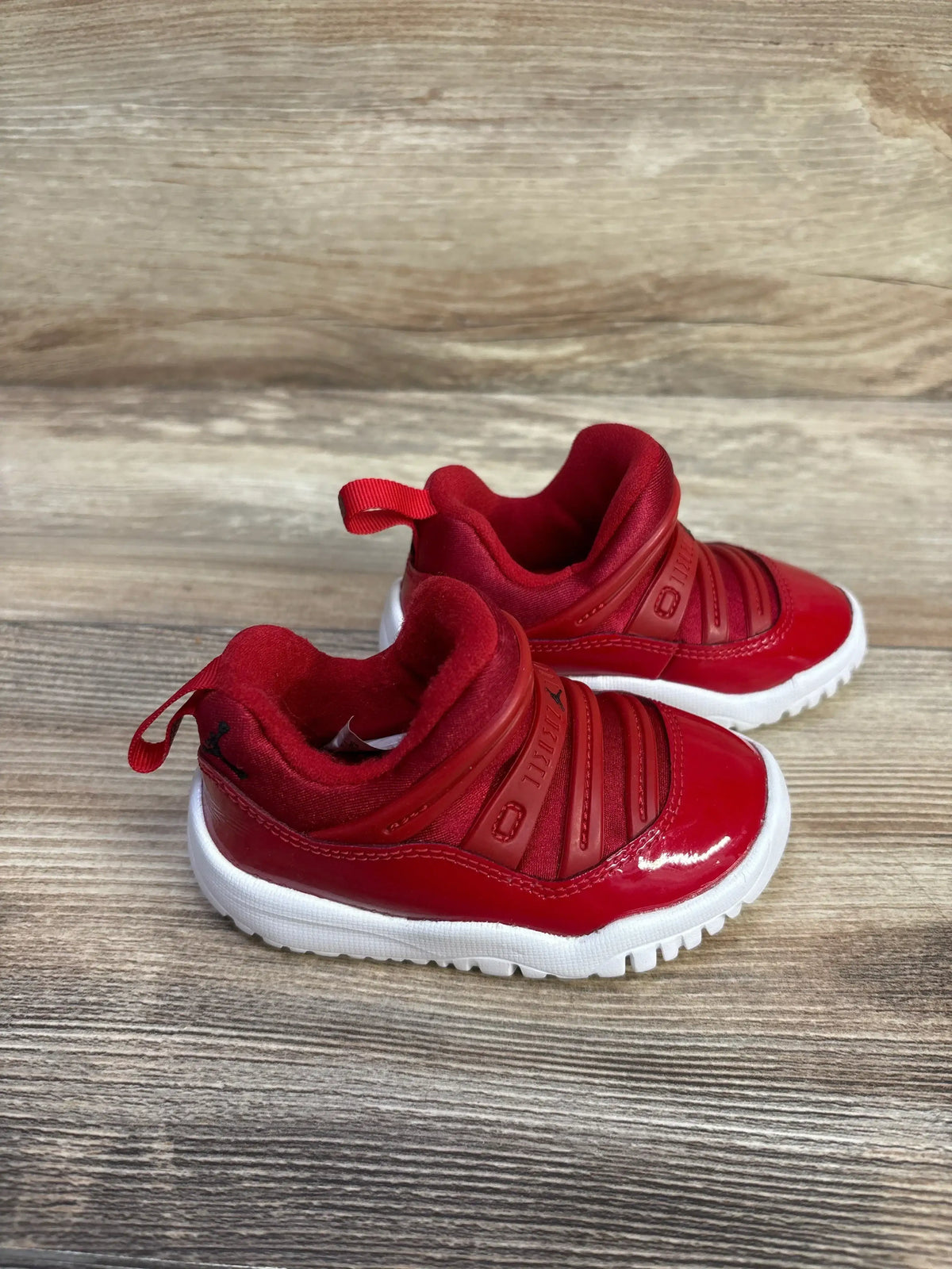 Angled view of Air Jordan 11 Retro Little Flex TD Gym Red sneakers with slip-on closure and white soles on wood flooring