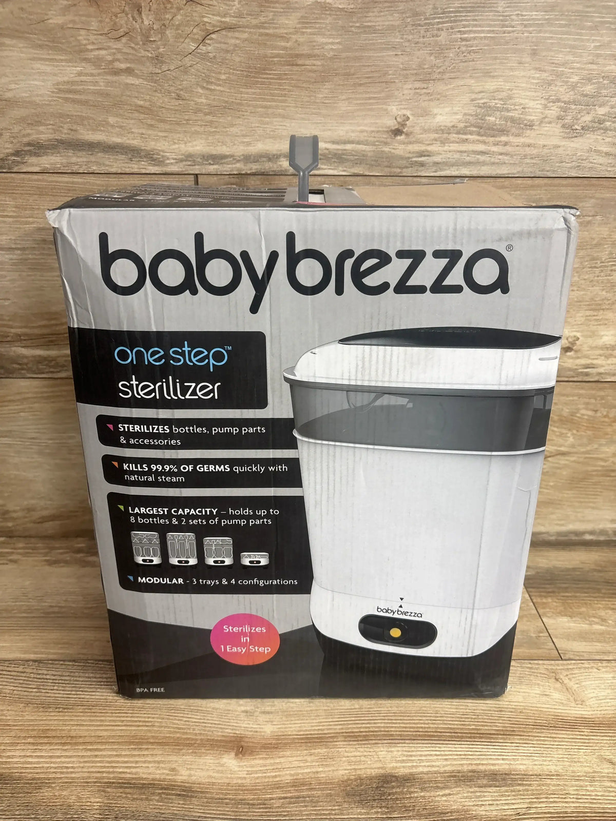 Front view of boxed Baby Brezza One Step sterilizer showing product image and feature callouts on wood background