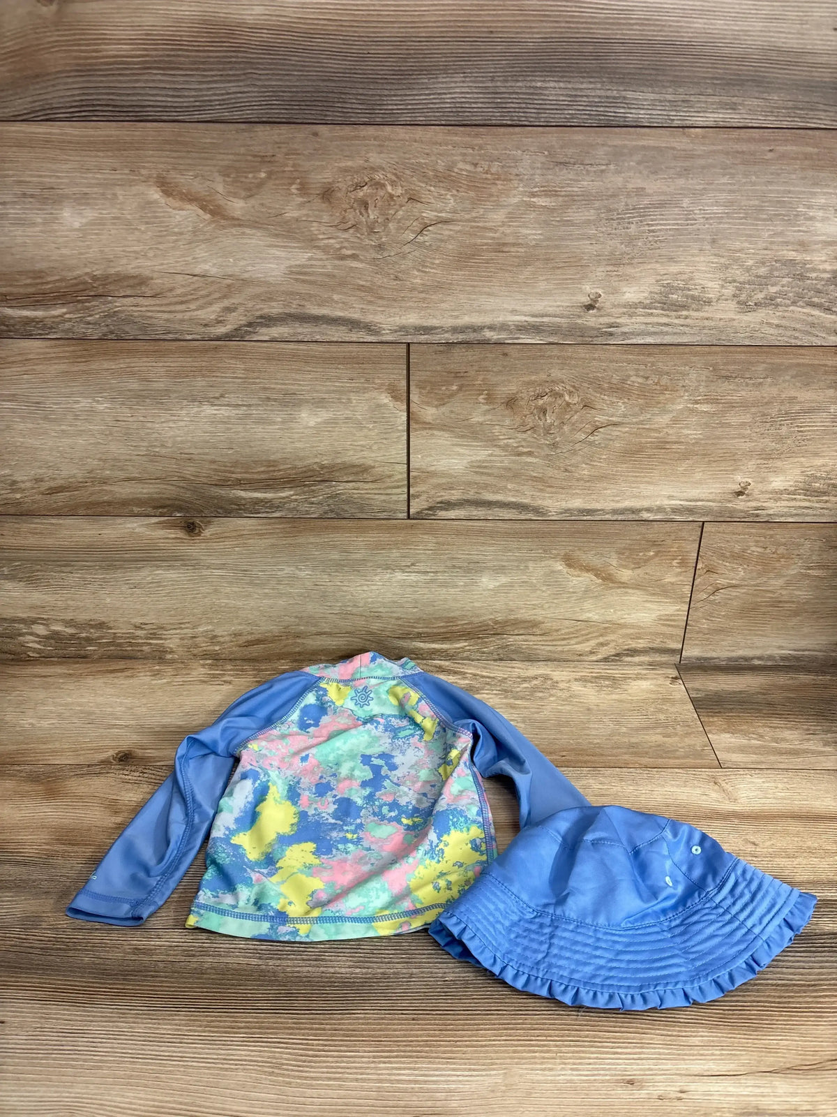 Top view of UV Skinz 2-piece blue rash guard shirt and bucket hat, size 3T, with colorful shirt front on wood floor
