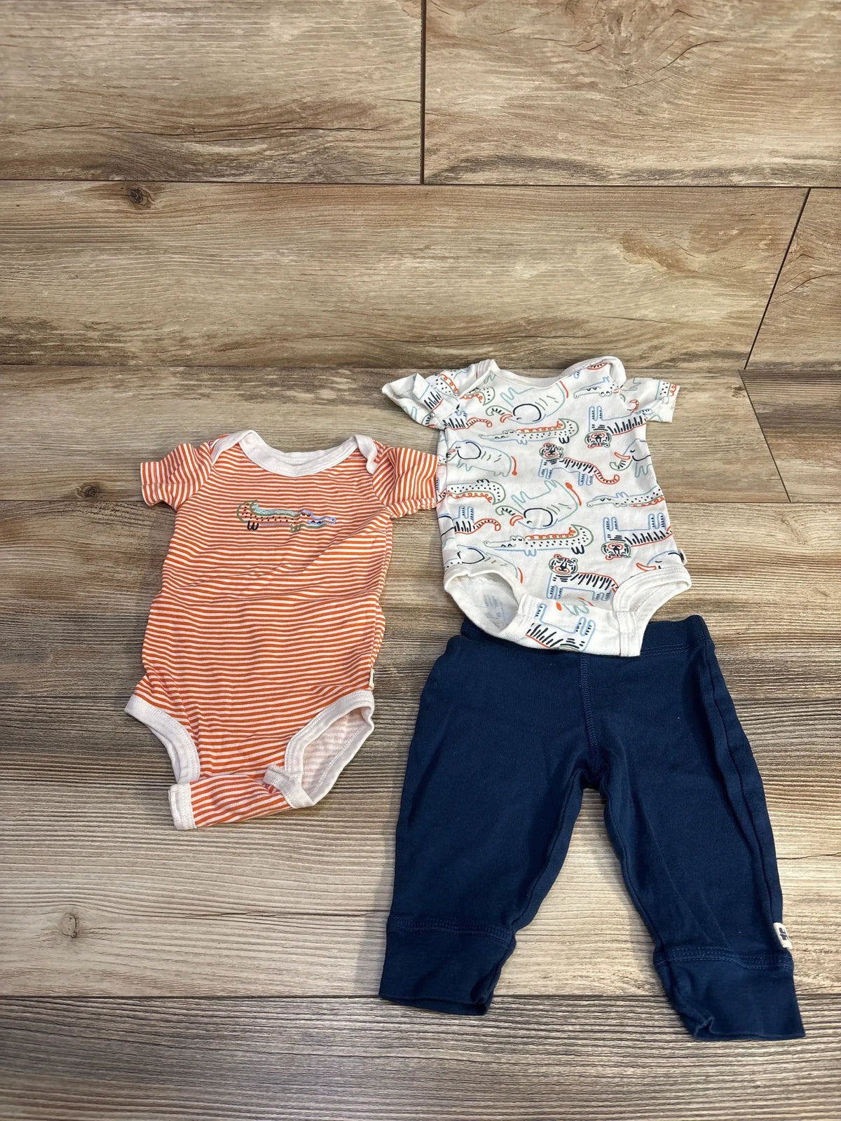 Flat lay of Huggies bodysuit set 3M showing orange striped and white animal print bodysuits with navy pants on wood floor