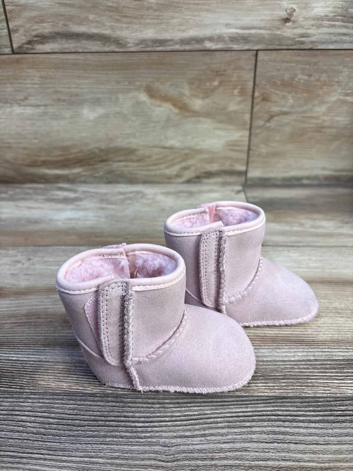Angled view of UGG Baby Classic soft sole pink booties showcasing suede exterior and plush pink lining on wooden background