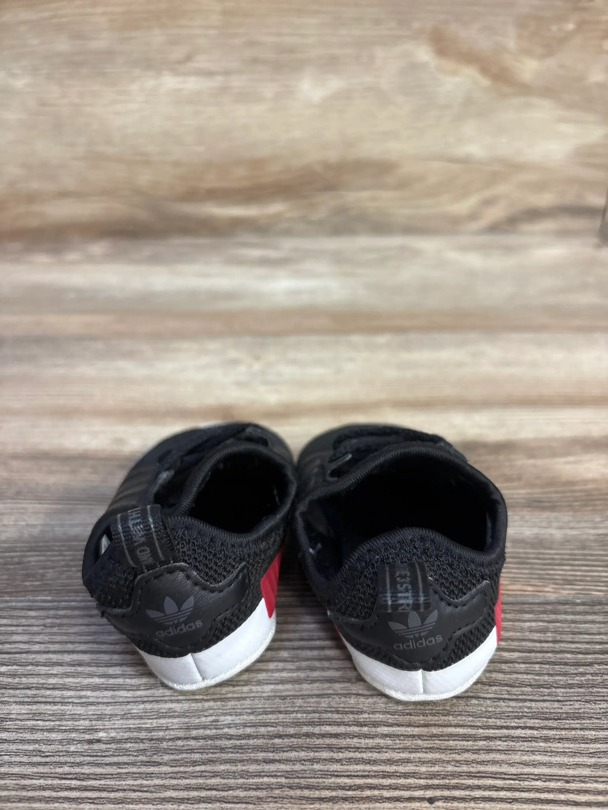Rear view of black Adidas NMD soft sole sneakers showcasing red accents and white outsoles on a wood-grain floor