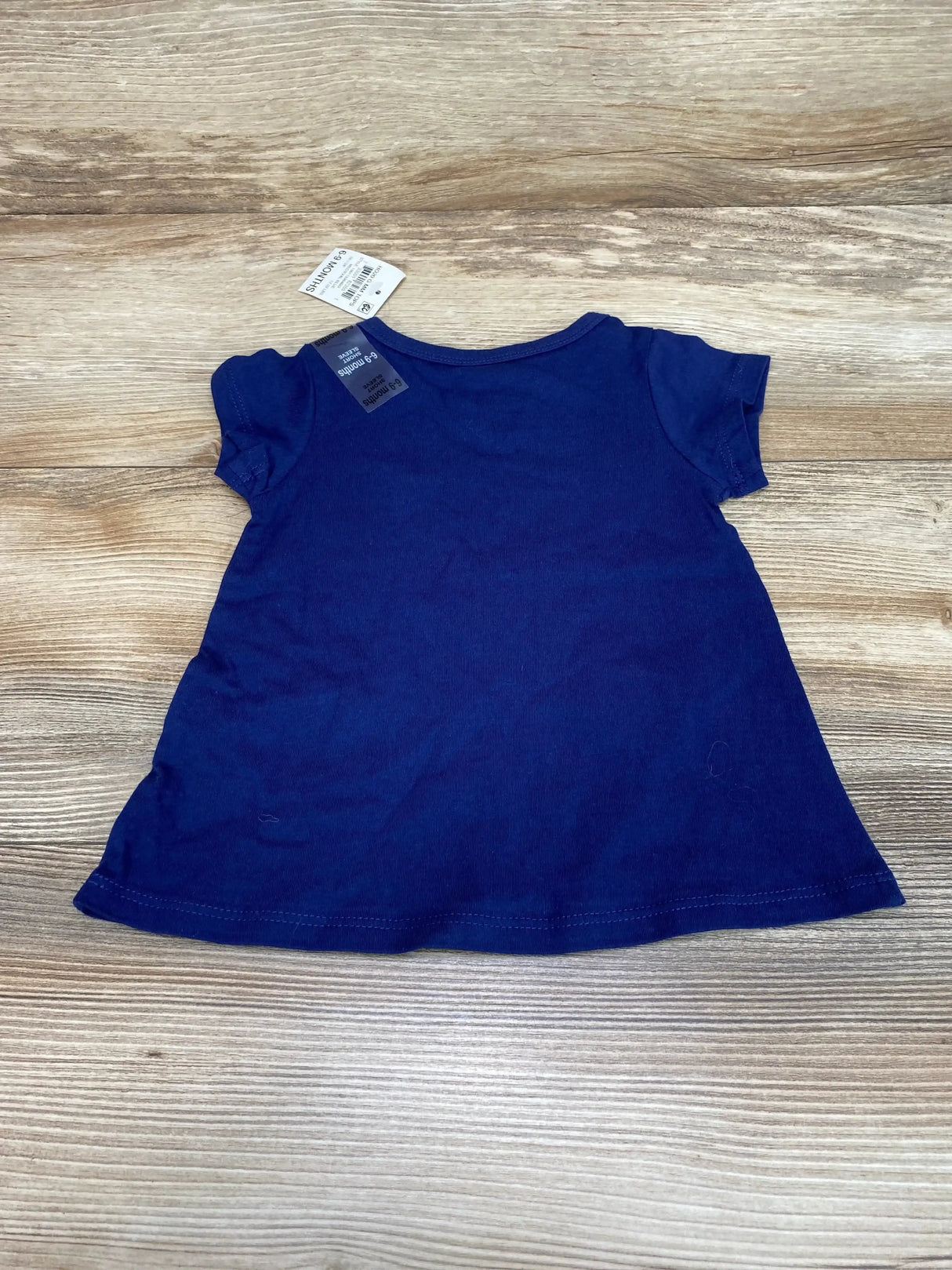 Flat-lay back of First Impressions navy baby tee showcasing short sleeves and attached 6-9M size tag on wood background