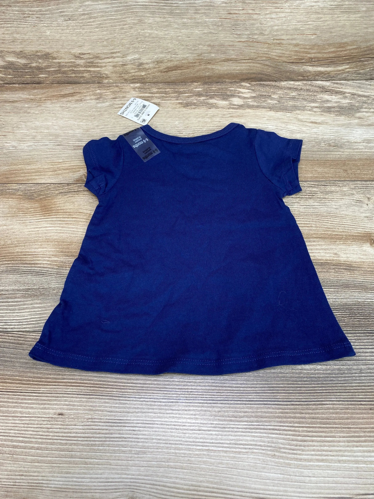 Flat-lay back of First Impressions navy baby tee showcasing short sleeves and attached 6-9M size tag on wood background