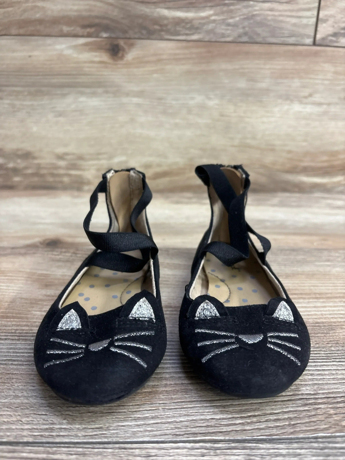 Pair of black children's ballet flats with silver cat face embroidery and ankle straps on wooden floor