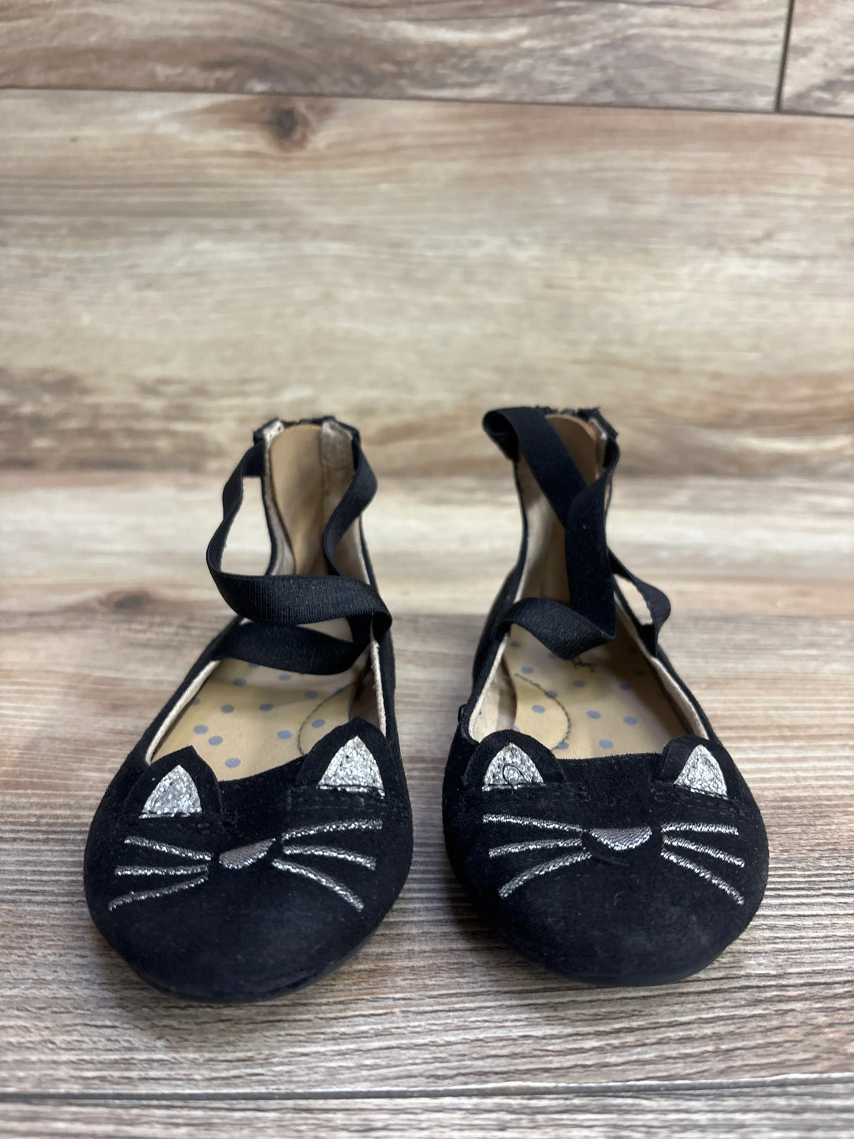 Pair of black children's ballet flats with silver cat face embroidery and ankle straps on wooden floor