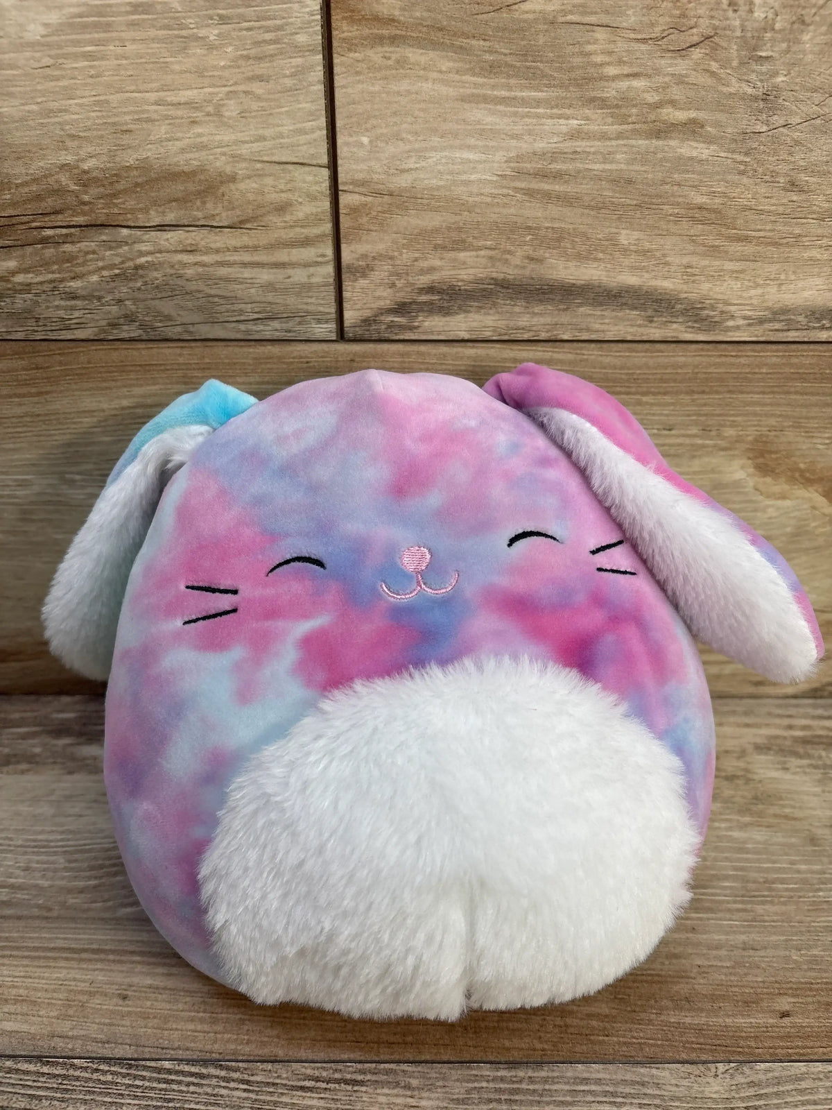 Front of 8in Ryder Bunny Squishmallow with pastel pink-blue plush, embroidered face and white fuzzy belly on wood