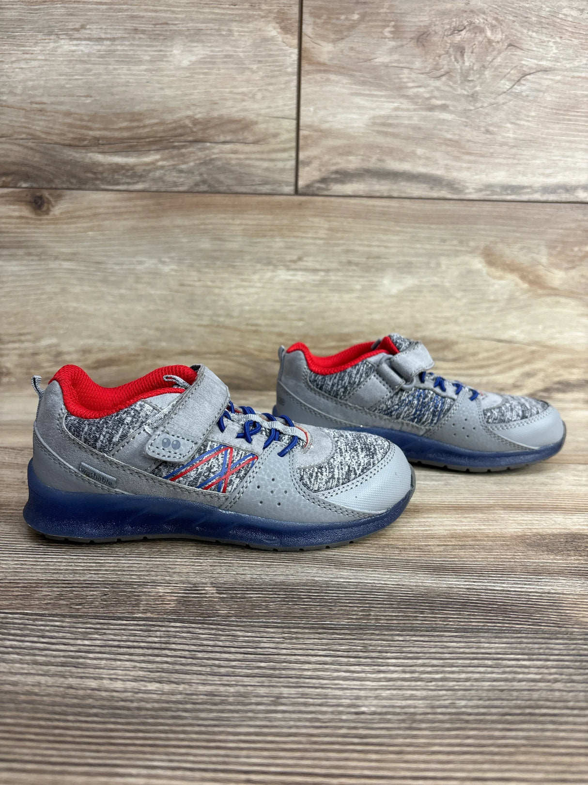 Pair of gray and blue children's sneakers with red inner lining and velcro straps on wooden floor