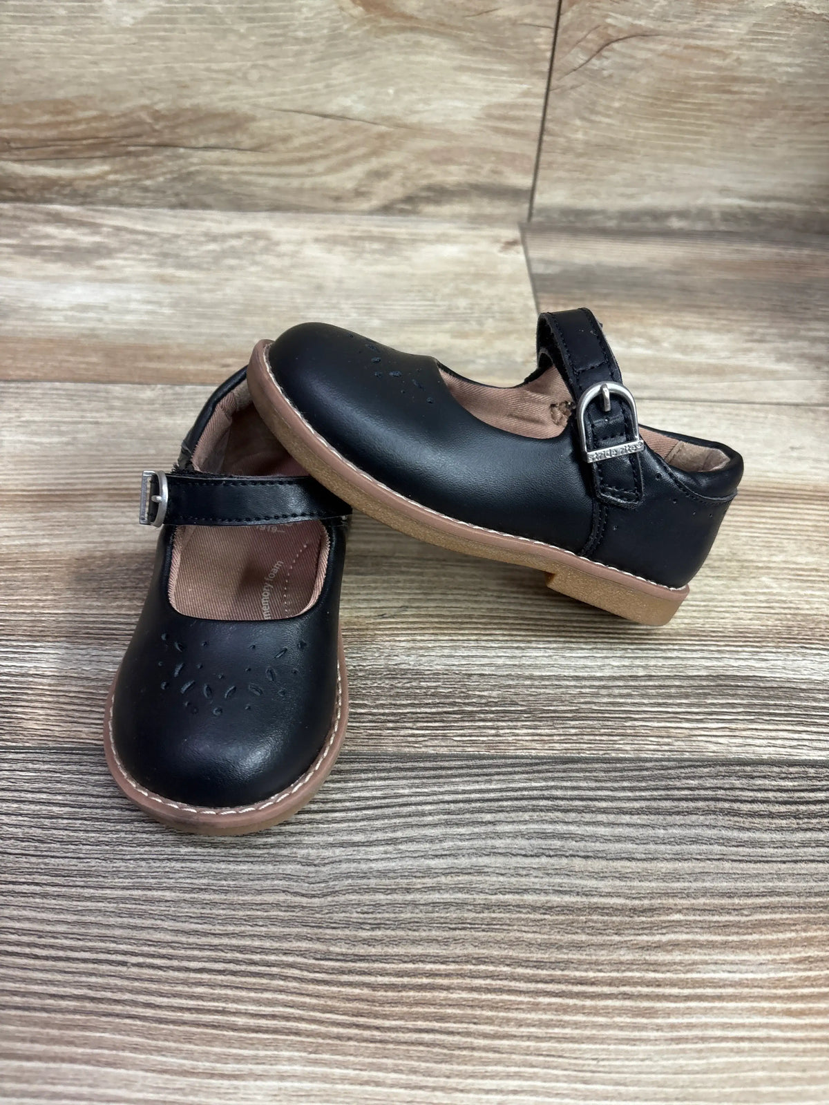 Top and side views of black Stride Rite Mara Mary Jane flats with buckle straps and perforated toes on a wood-look surface