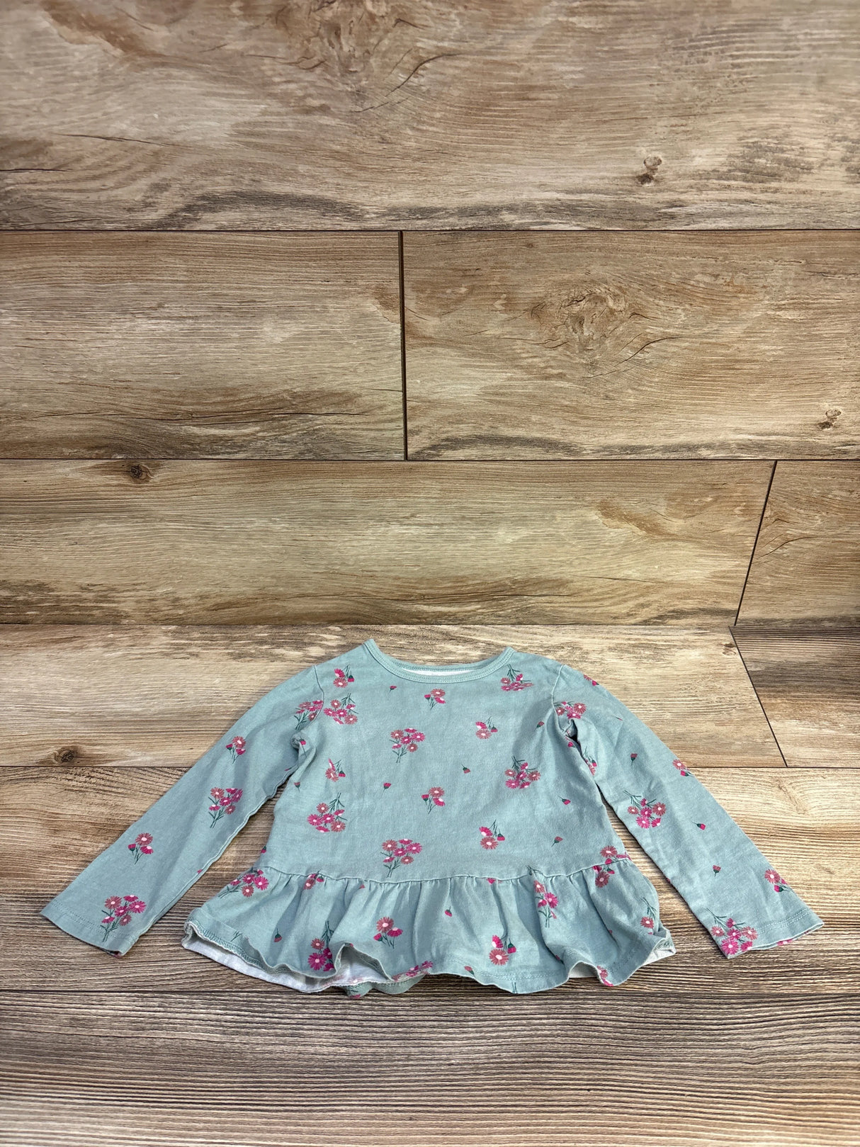 Flat lay of toddler peplum top, size 2T, showcasing teal-green fabric with pink floral print and ruffle hem on wood background
