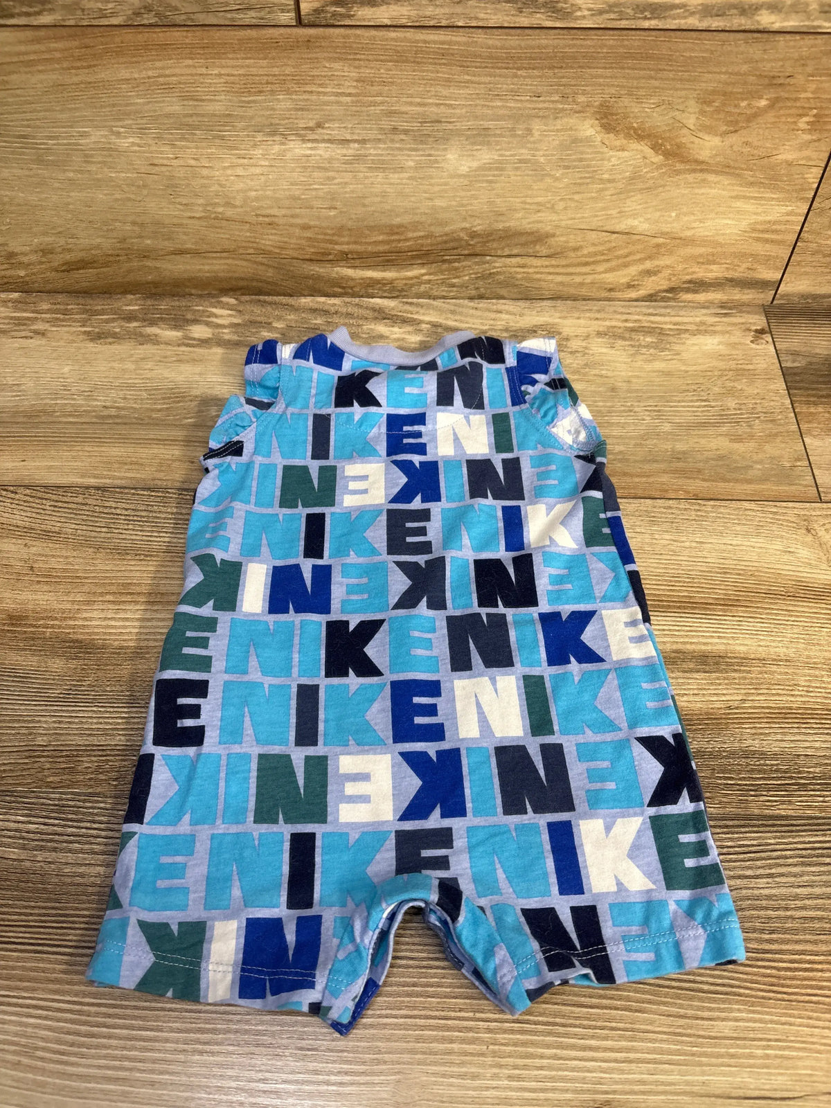 Front view of Nike blue flutter sleeve romper for 6 months showcasing a repeating multicolor Nike lettering pattern on a wood floor