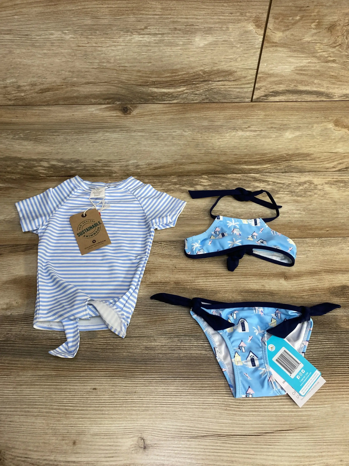Flat lay of toddler blue halter bikini with beach-house print, tie-side bottoms, and blue-and-white striped rash top on wood floor