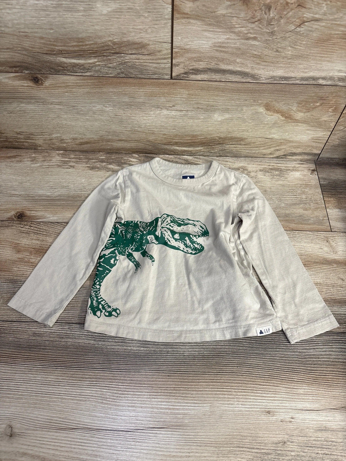 Beige long sleeve kids shirt with green dinosaur graphic, laid flat on wood floor