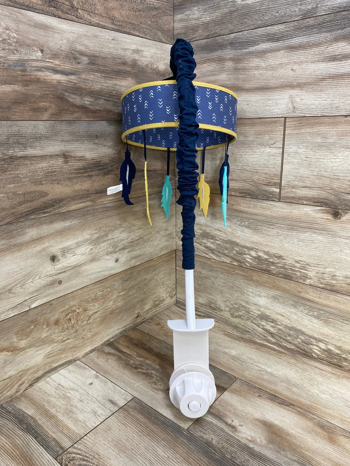 Front view of MY Baby Sam Crib Mobile showing navy drum with yellow trim and hanging blue, teal, and yellow felt feathers on wooden floor with white clamp mount