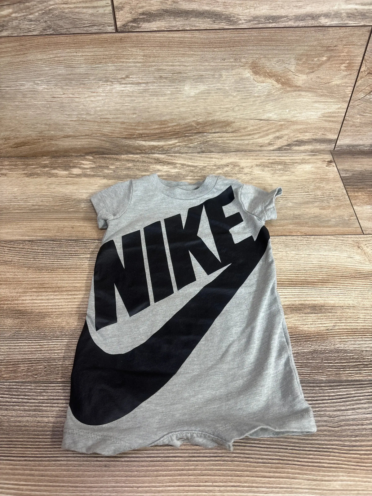 Flat lay front view of grey Nike shortie romper size 3M showing large black NIKE wordmark and swoosh on wood background