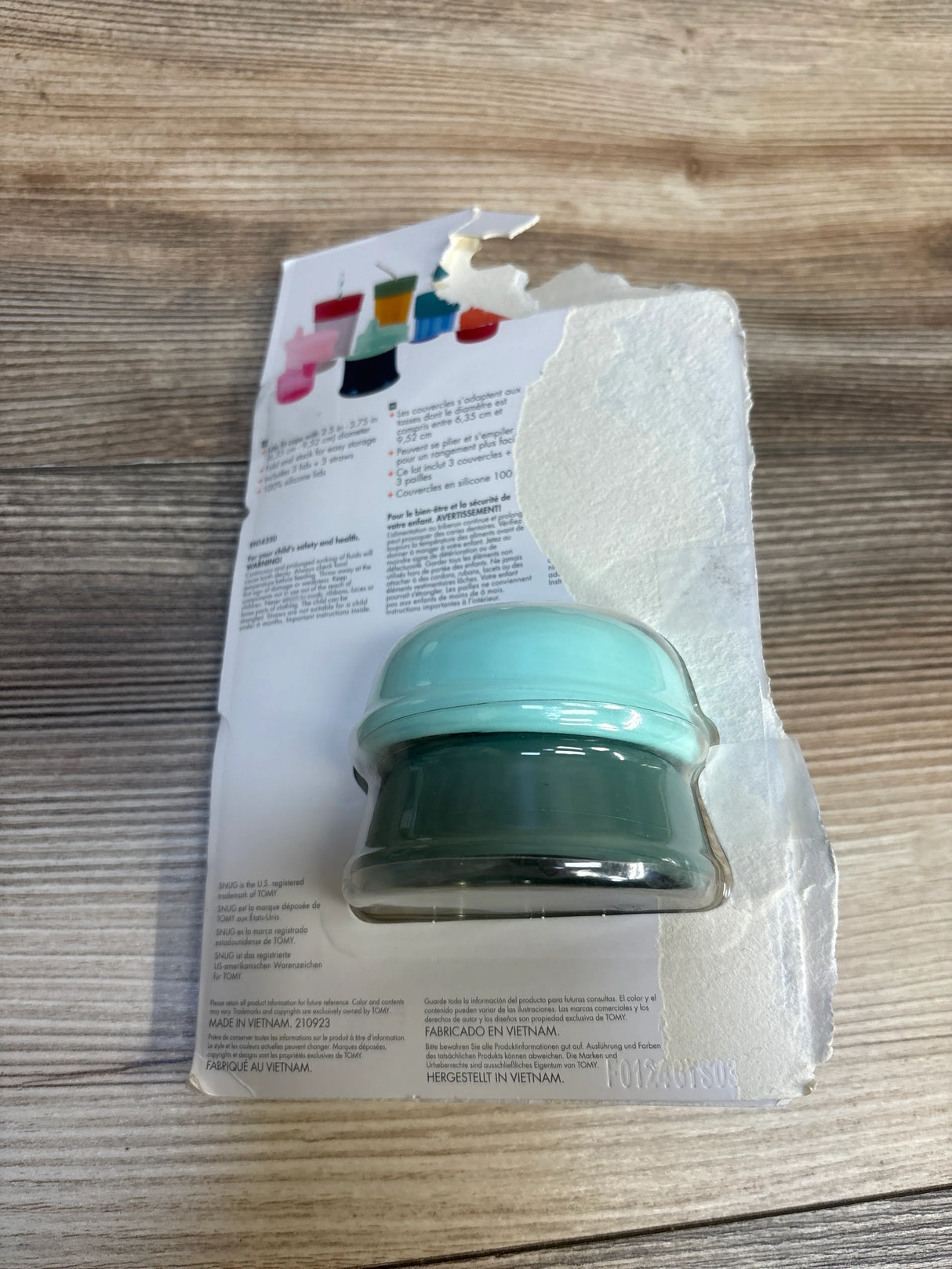 Two silicone sippy cup lids in green and aqua, packaged on torn cardboard backing on wood surface.