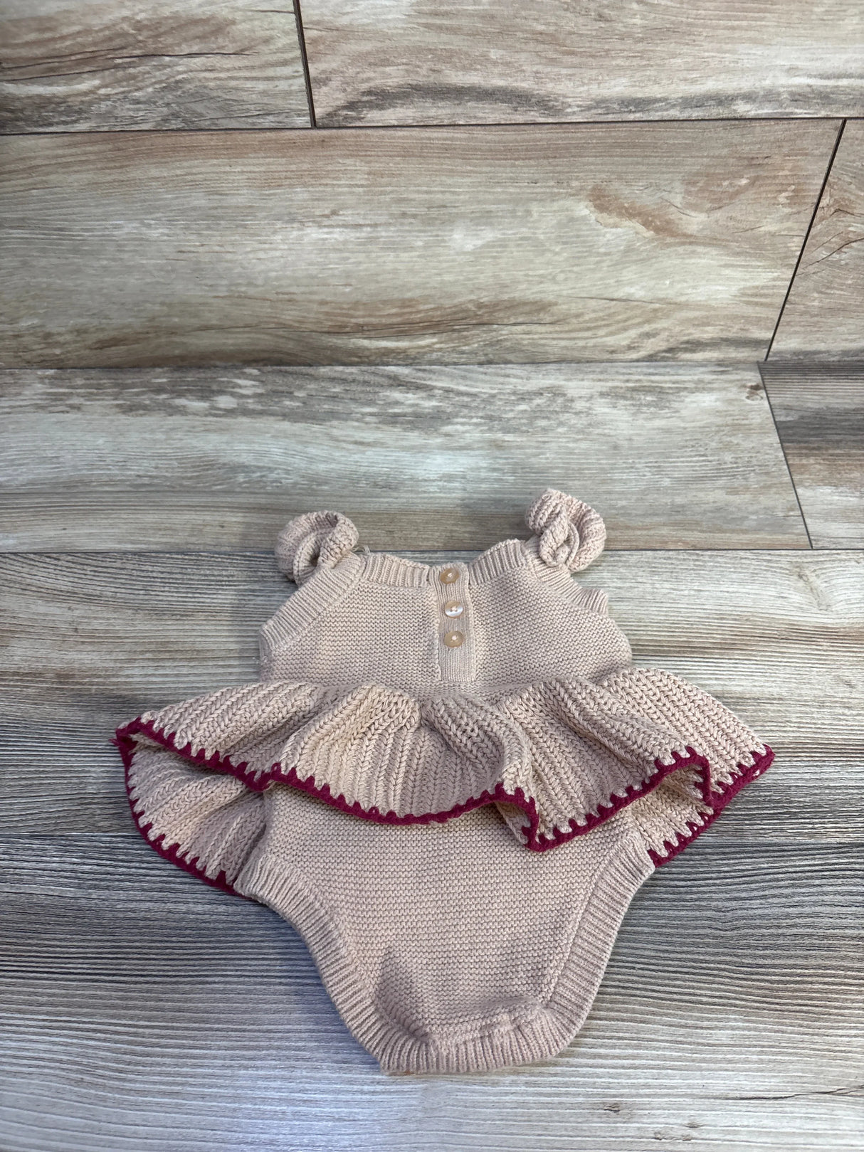 Flat lay of Nicole Miller knit skirted bodysuit size 3-6 months showcasing ruffled skirt with burgundy trim, knotted straps and buttons on wood background