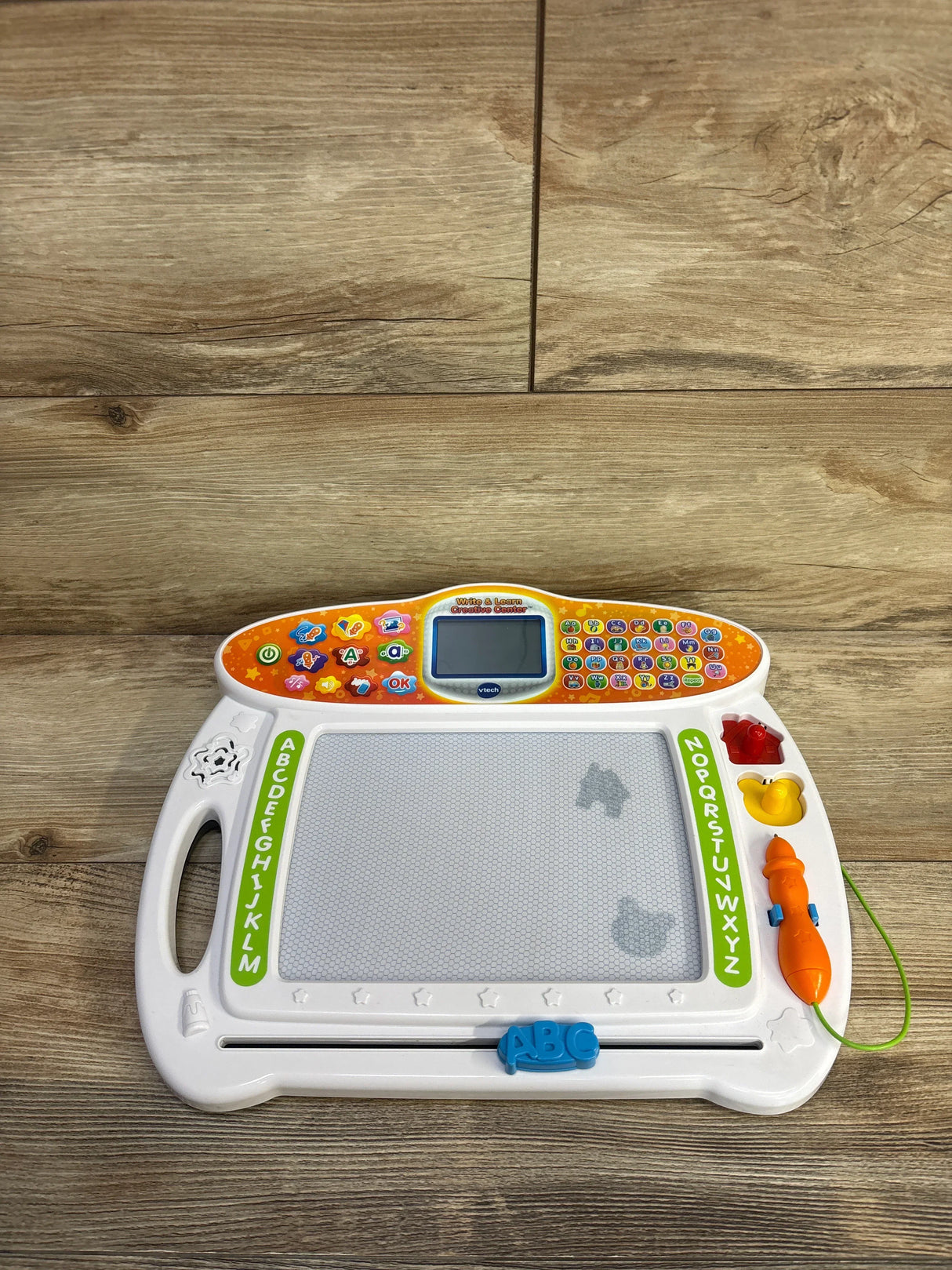 Front of VTech Write and Learn Creative Center with magnetic drawing board, attached stylus, alphabet stamps on wood floor
