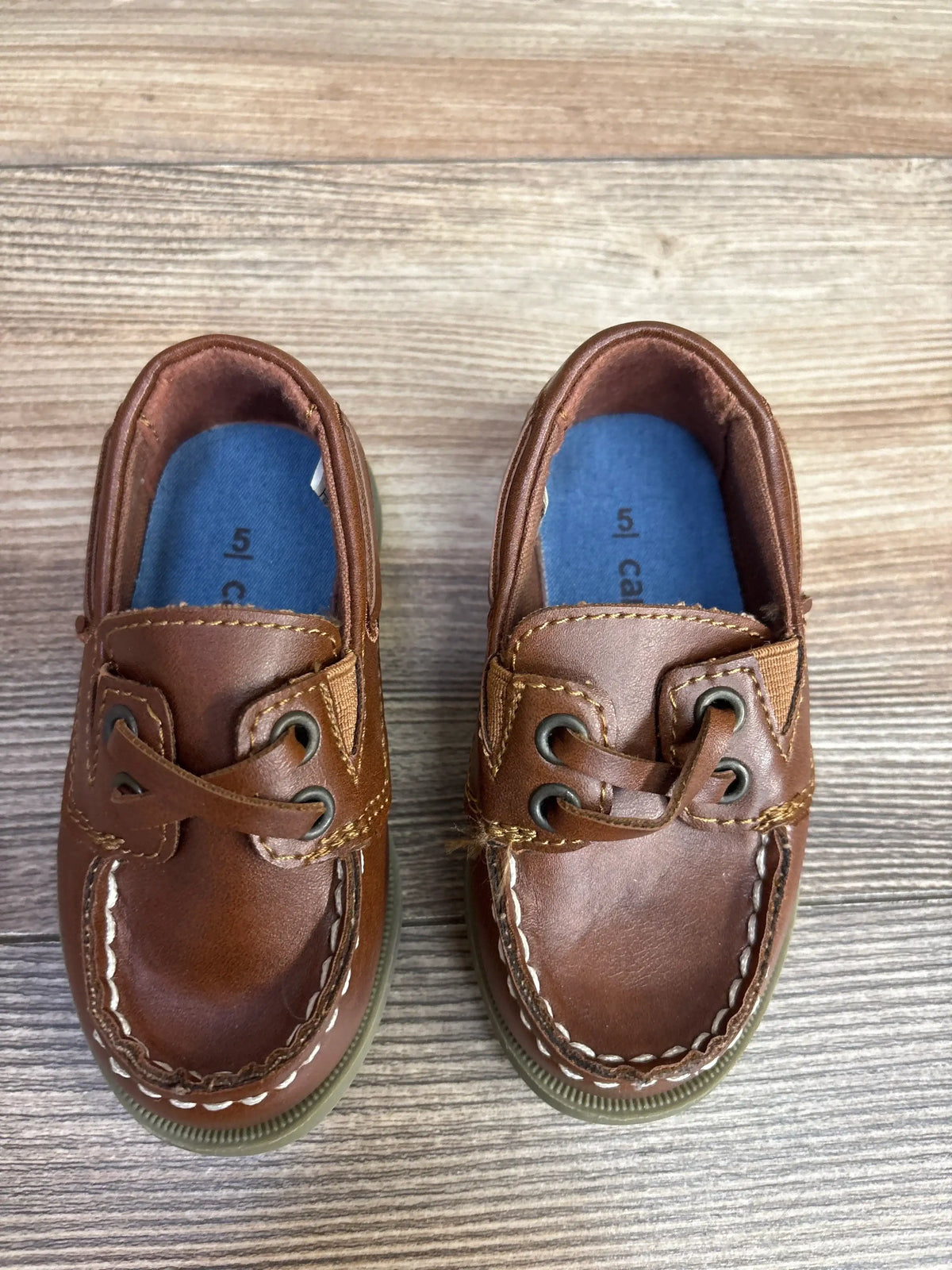 Top-down of Carter’s Bauk slip-on brown leather toddler boat shoes size 5C showing white stitching, blue insoles on wood floor