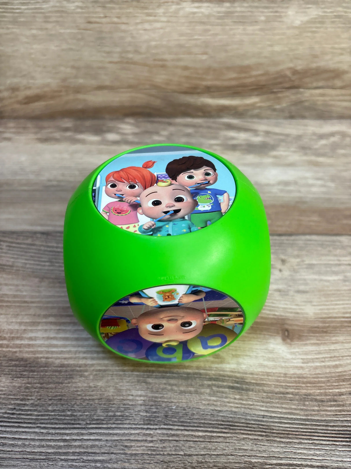 Top view of Cocomelon Tumble Tunes musical toy showing green rounded block with character stickers on a wooden surface