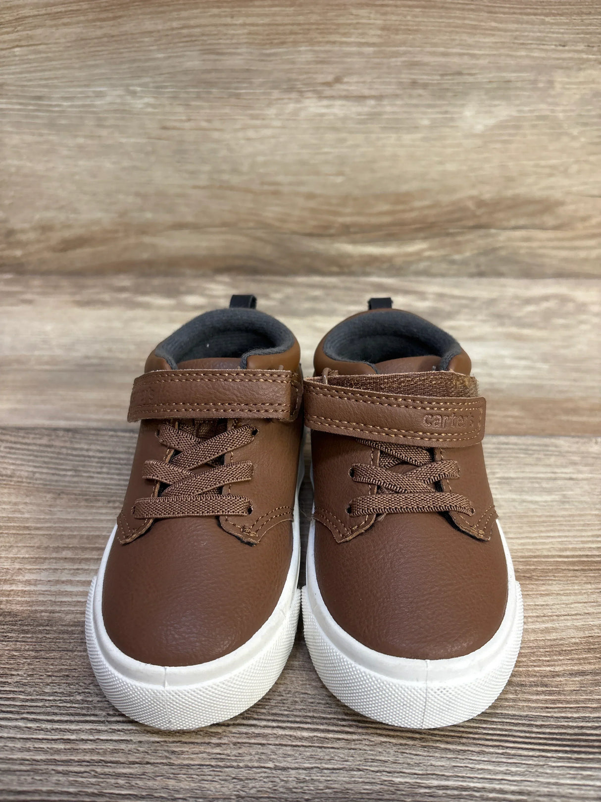 Front view of Carter's brown toddler sneakers with Velcro strap, faux laces and white rubber sole on wood-grain surface