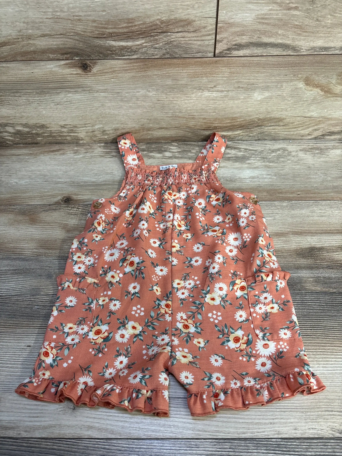 Flat lay of Nicole Miller coral floral shortall showcasing smocked bodice, ruffle hem, and front pockets on a wood background