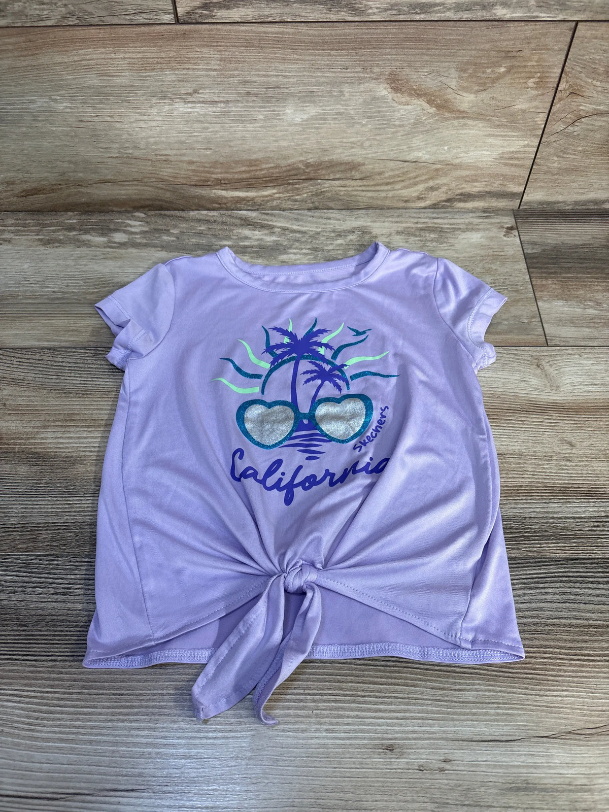 Front view of Skechers California shirt showcasing purple short sleeves, palm tree and sunglasses graphic, and front tie on a wood floor