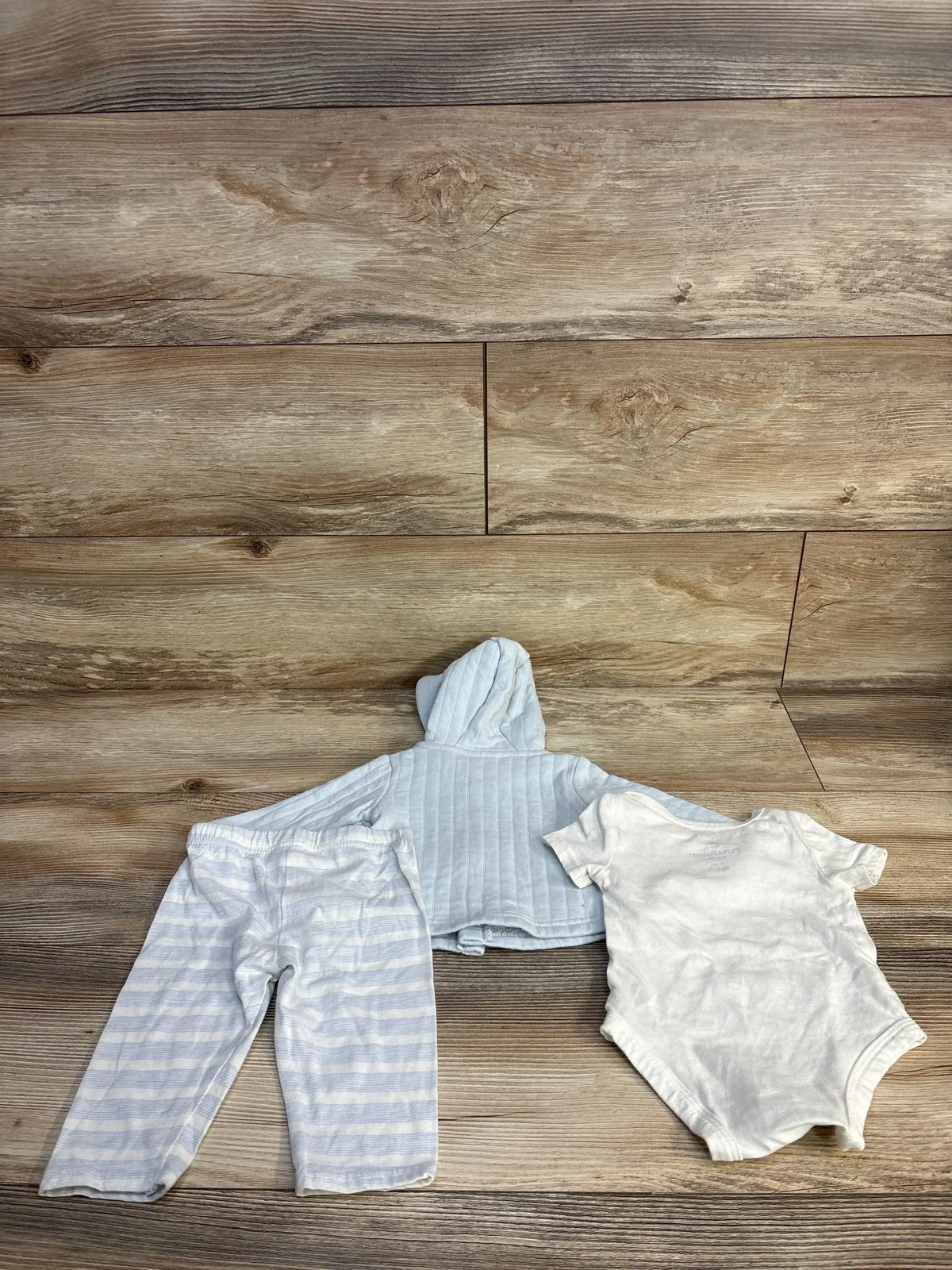 Flat lay of Peter Rabbit 3-piece baby outfit: blue hooded jacket, striped pants, and white bodysuit on wood floor