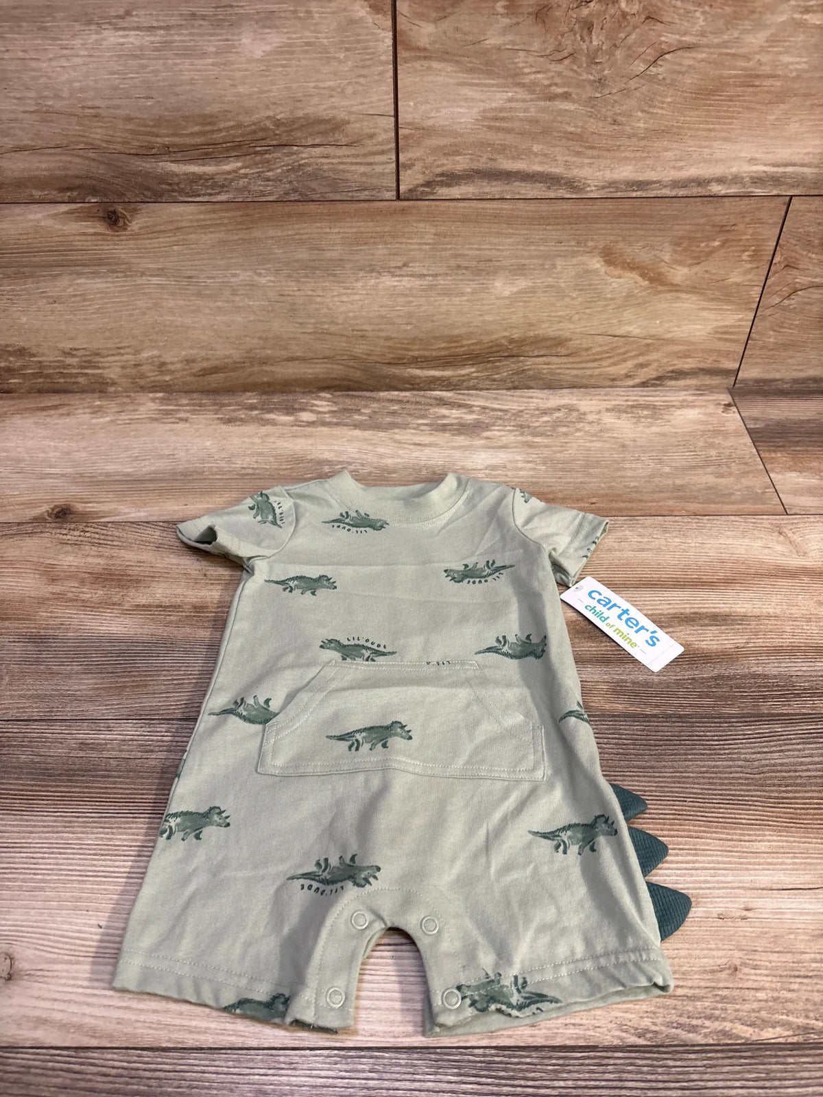 Flat lay of sage green shortie romper with dinosaur print, front pocket, snap crotch and fabric spikes on wood background