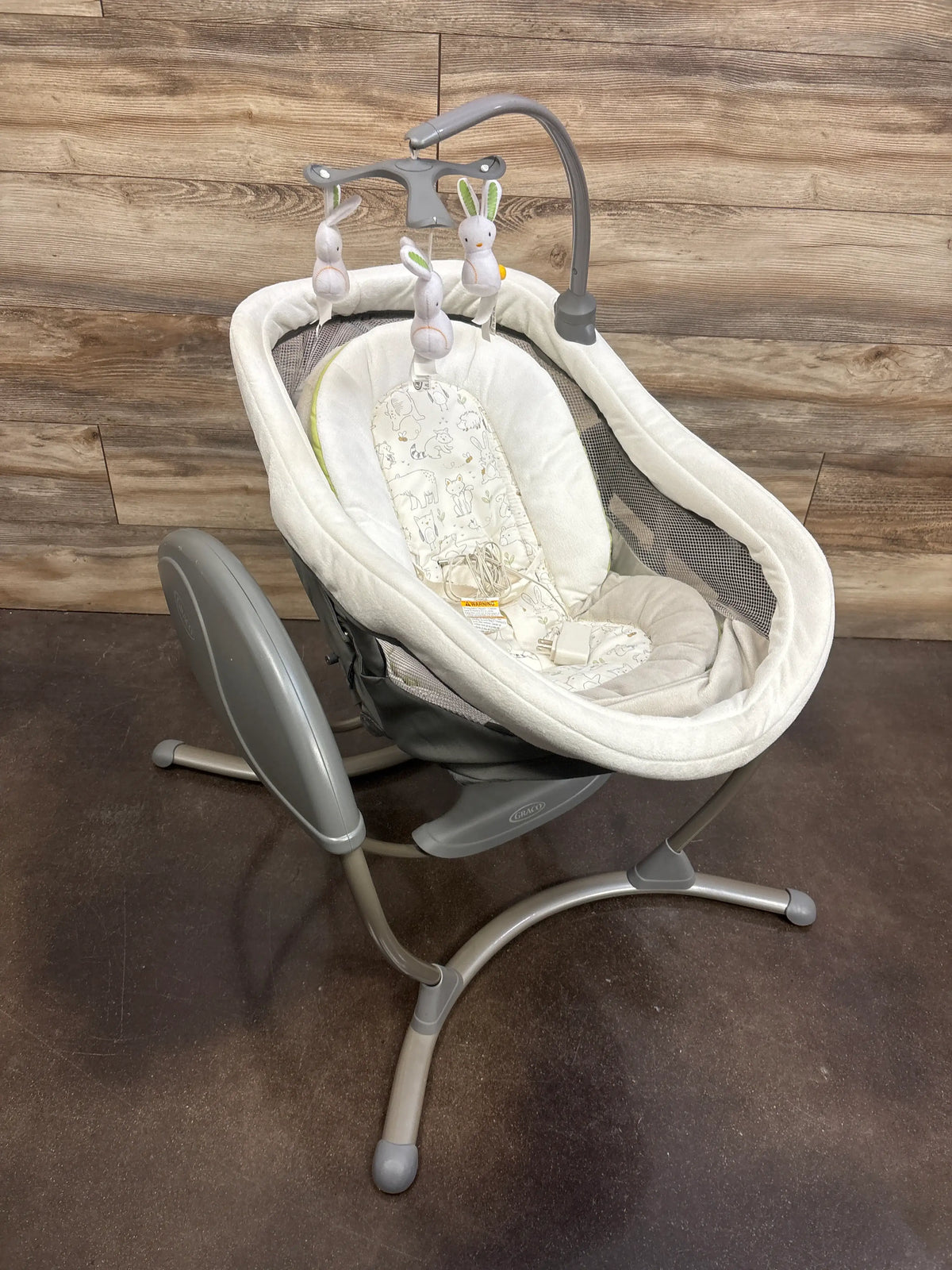 Angled view of Graco DuoGlider gliding swing showcasing white seat, mesh sides, toy bar, and gray frame on wood floor