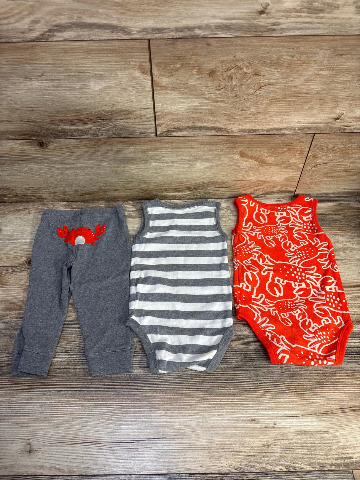 Flat lay of Carter's 3-piece baby set: grey pants with red crab, grey-striped tank, and red crab-print bodysuit on wood