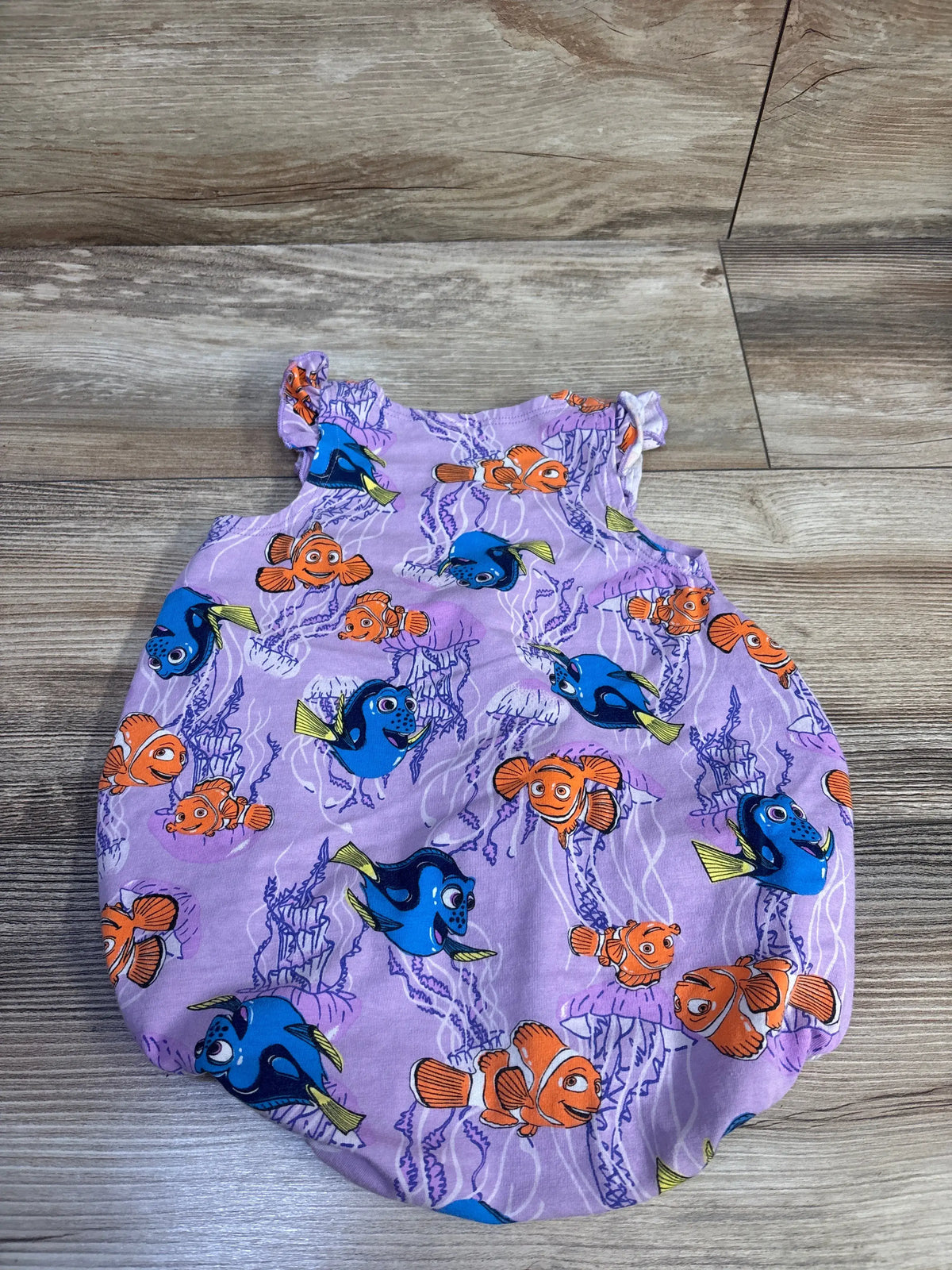 Flat lay of a purple Bonds x Disney Finding Nemo bubble romper showcasing Nemo and Dory character print on a wood-grain floor