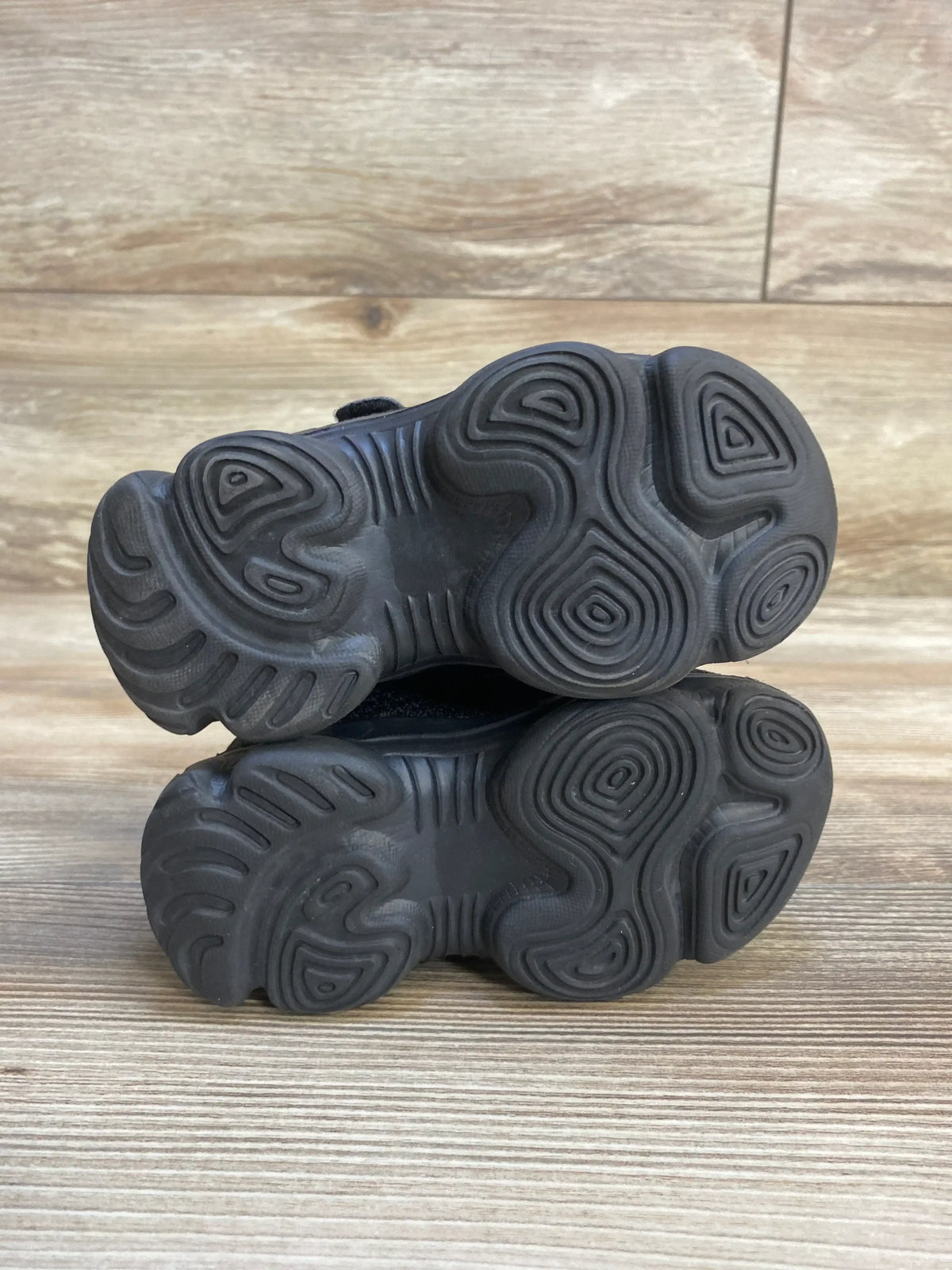 Pair of black kids' sneakers with textured rubber soles on wood surface
