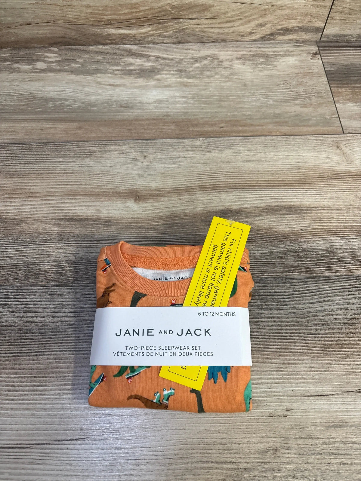 Top down view of folded orange Janie and Jack two piece pajama set showcasing skate riding dinosaur print and product band on wood floor