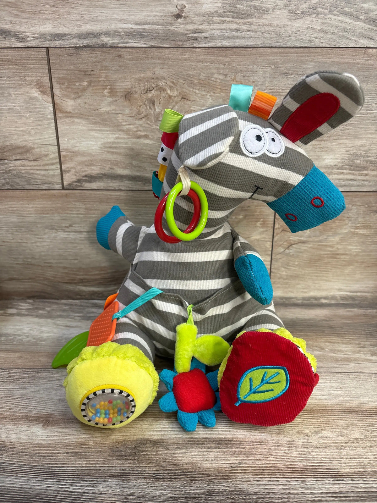 Front view of Dolce Zeddy The Zebra Activity Plush showcasing striped zebra design and attached textured toys on a wood floor