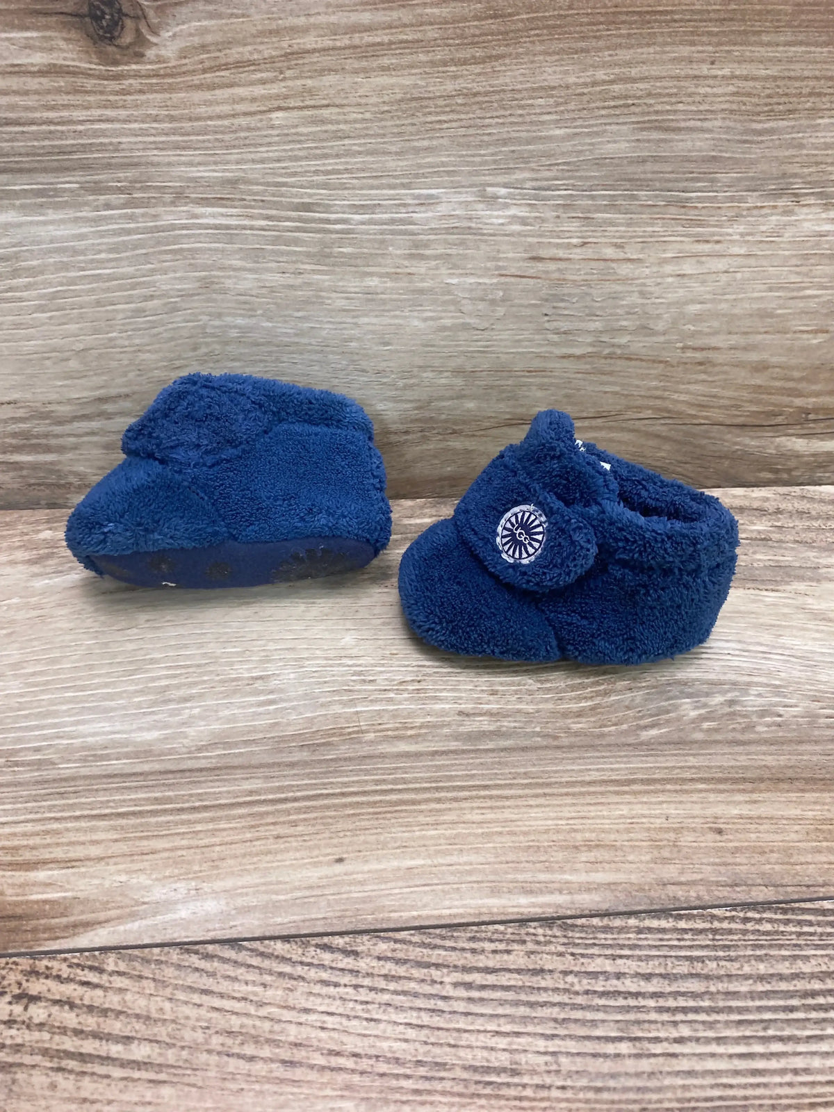 Side and front view of UGG Baby Bixbee boot in blue showing plush fleece and Velcro strap with logo on wooden background