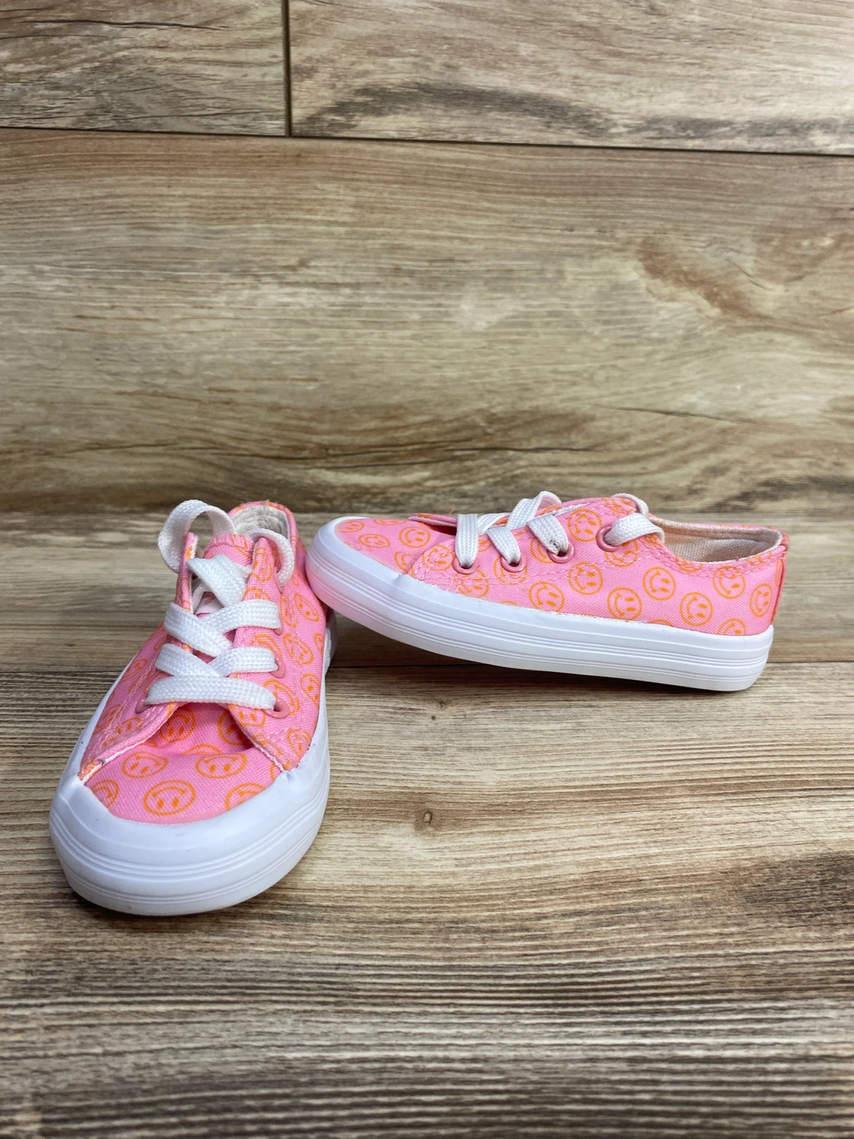 Pair of pink toddler canvas sneakers with orange smiley faces and white laces on wooden surface